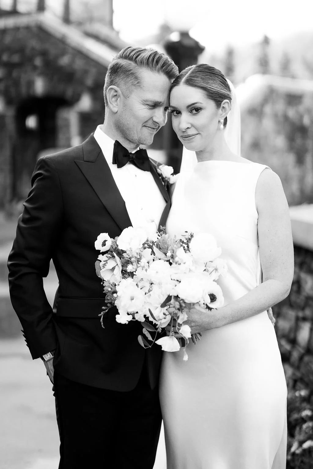 Black and white photo of a bride and groom on their wedding day, standing outdoors, with the groom wearing a tuxedo and the bride in a sleeveless white dress, holding a bouquet of flowers.