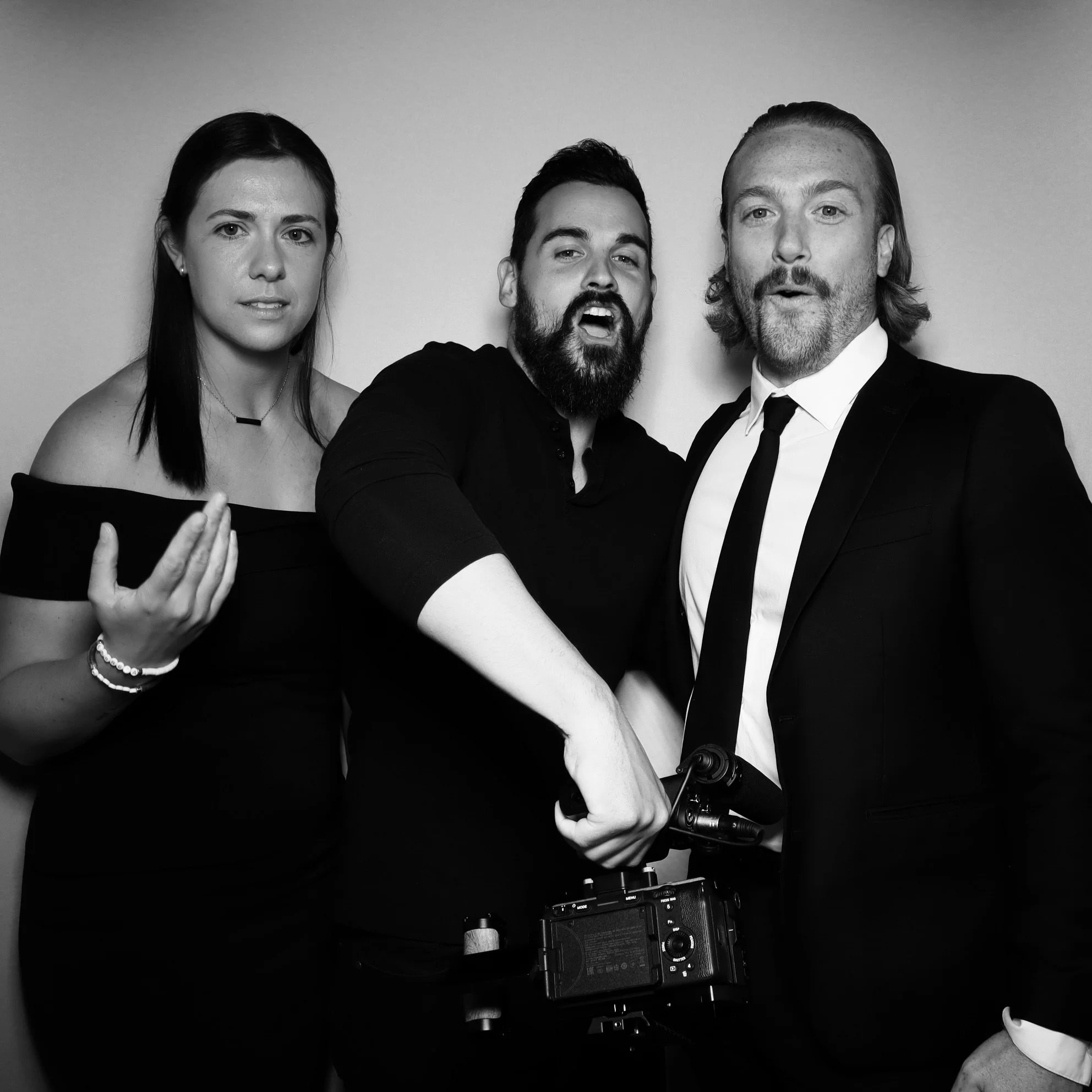 Black and white photo of three people, a woman on the left, a man in the middle, and a man on the right, posing together in formal attire, with the man in the middle holding a camera.