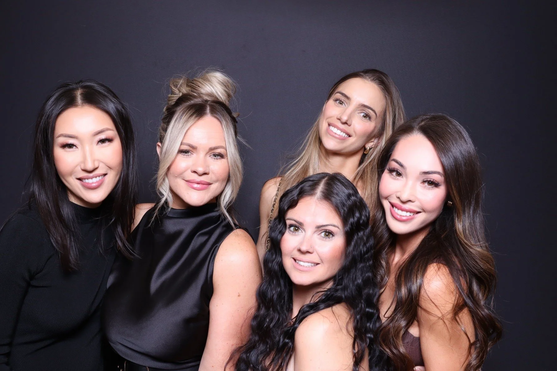 Six women smiling and posing together against a dark background, with diverse hairstyles and hair colors.