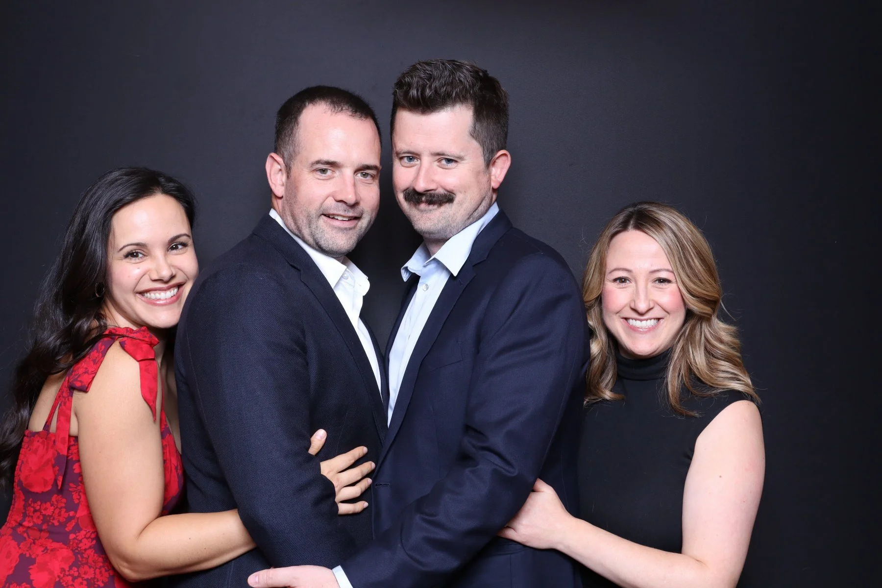 Four people standing close together, two men in suits in the center and two women on either side, smiling against a black background.