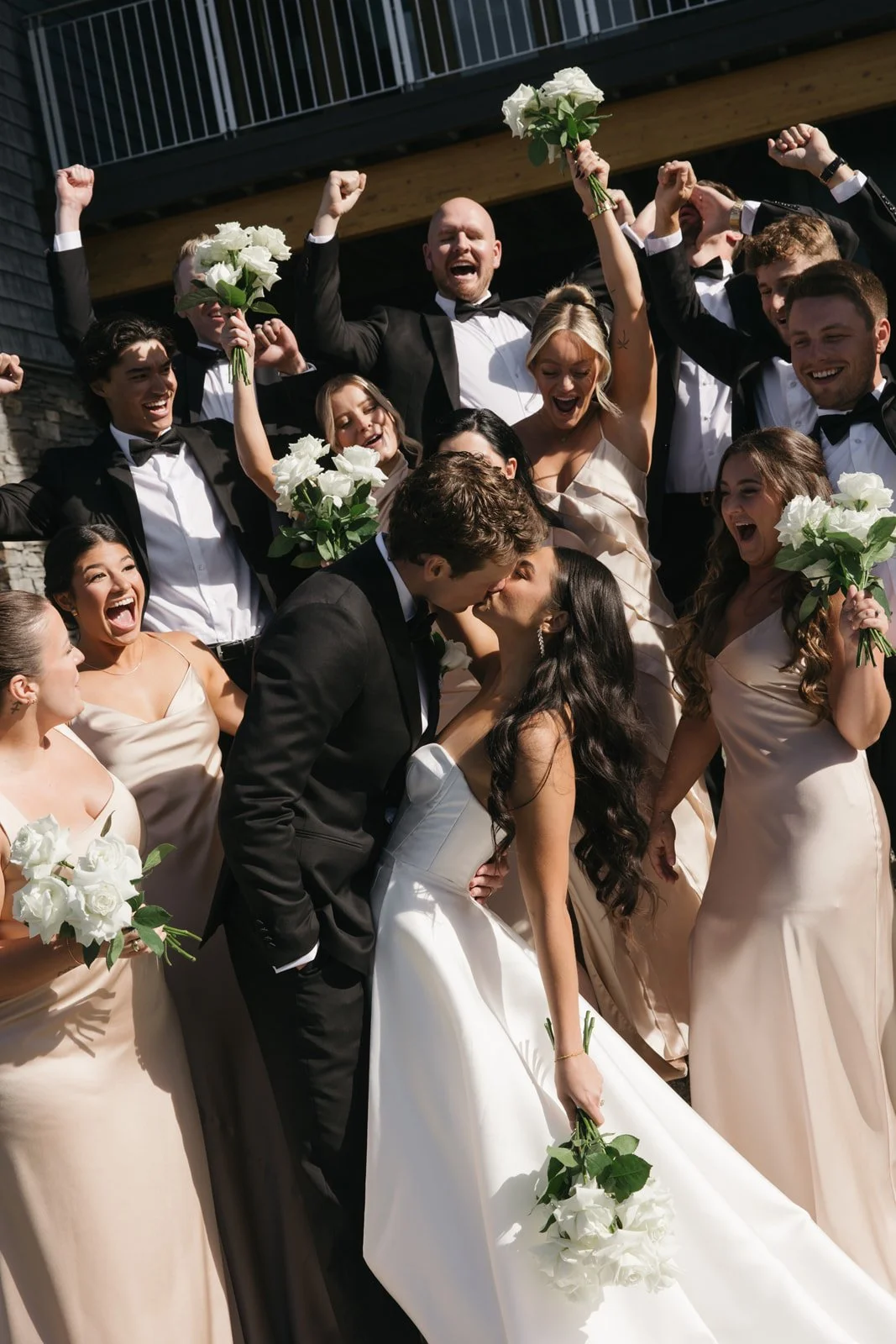 A wedding celebration with the bride and groom kissing, surrounded by their wedding party of bridesmaids and groomsmen cheering and holding white bouquets.