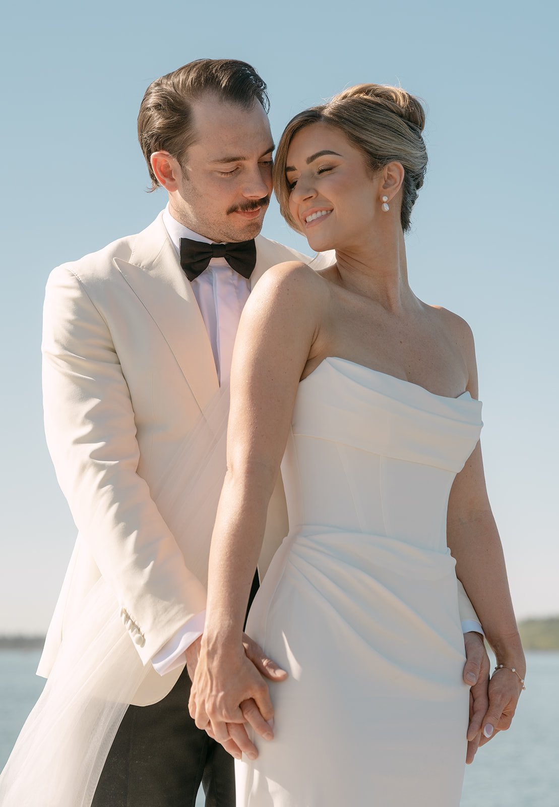 A bride and groom standing close together outdoors, holding hands, with the ocean and blue sky in the background. The groom is in a white tuxedo with a black bow tie, and the bride is wearing a strapless white wedding gown. They are smiling and embra