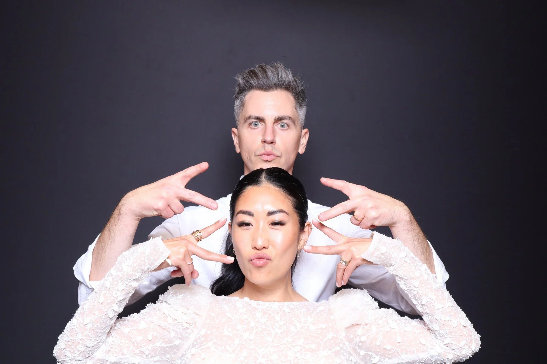 A man and woman posing playfully against a dark background. The man stands behind the woman, making peace signs near his face, and the woman puckers her lips while winking, with her hands near her face.