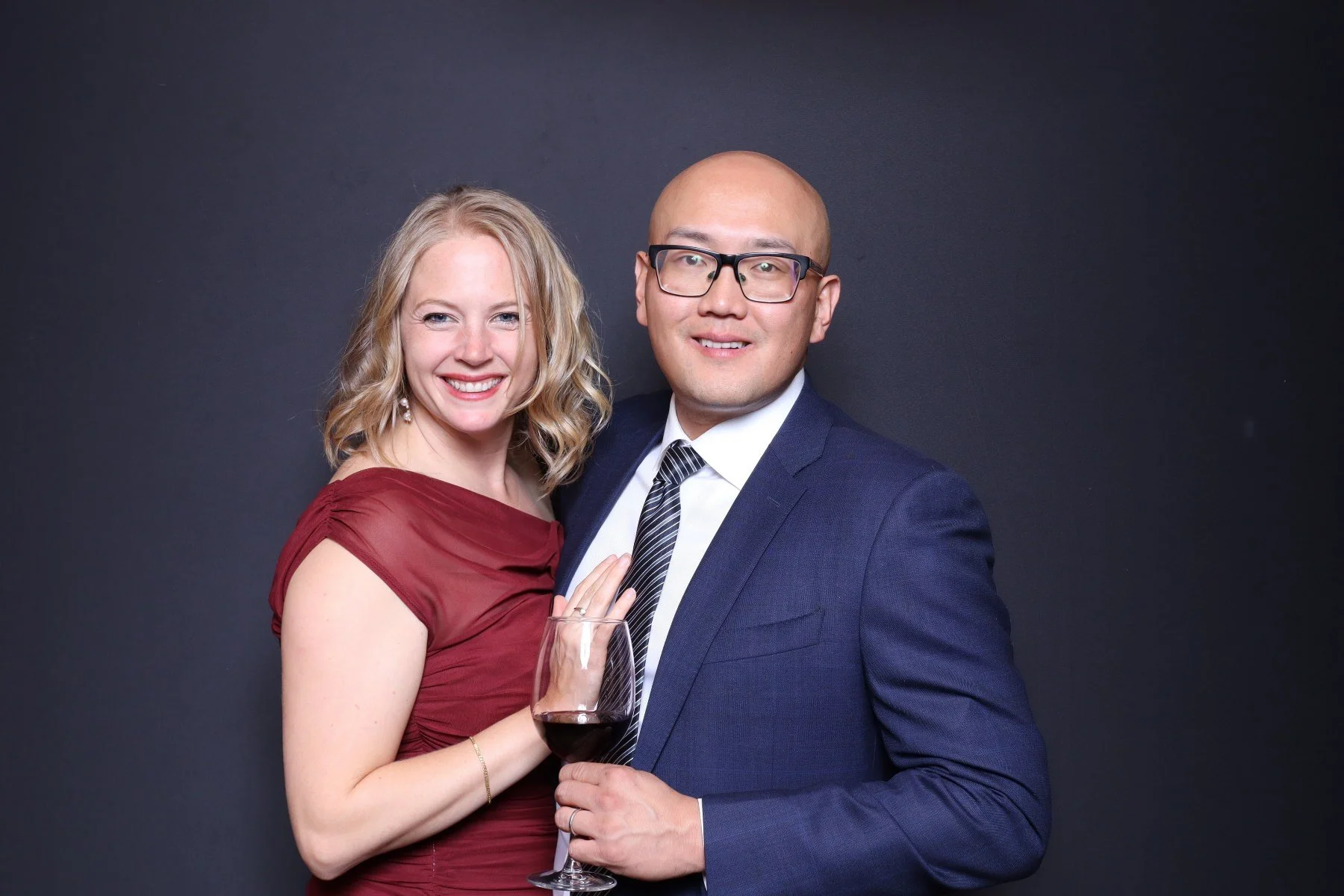 A couple dressed in formal attire, smiling at the camera; the woman has blonde curly hair and is holding a glass of red wine, while the man is bald, wearing glasses and a blue suit.