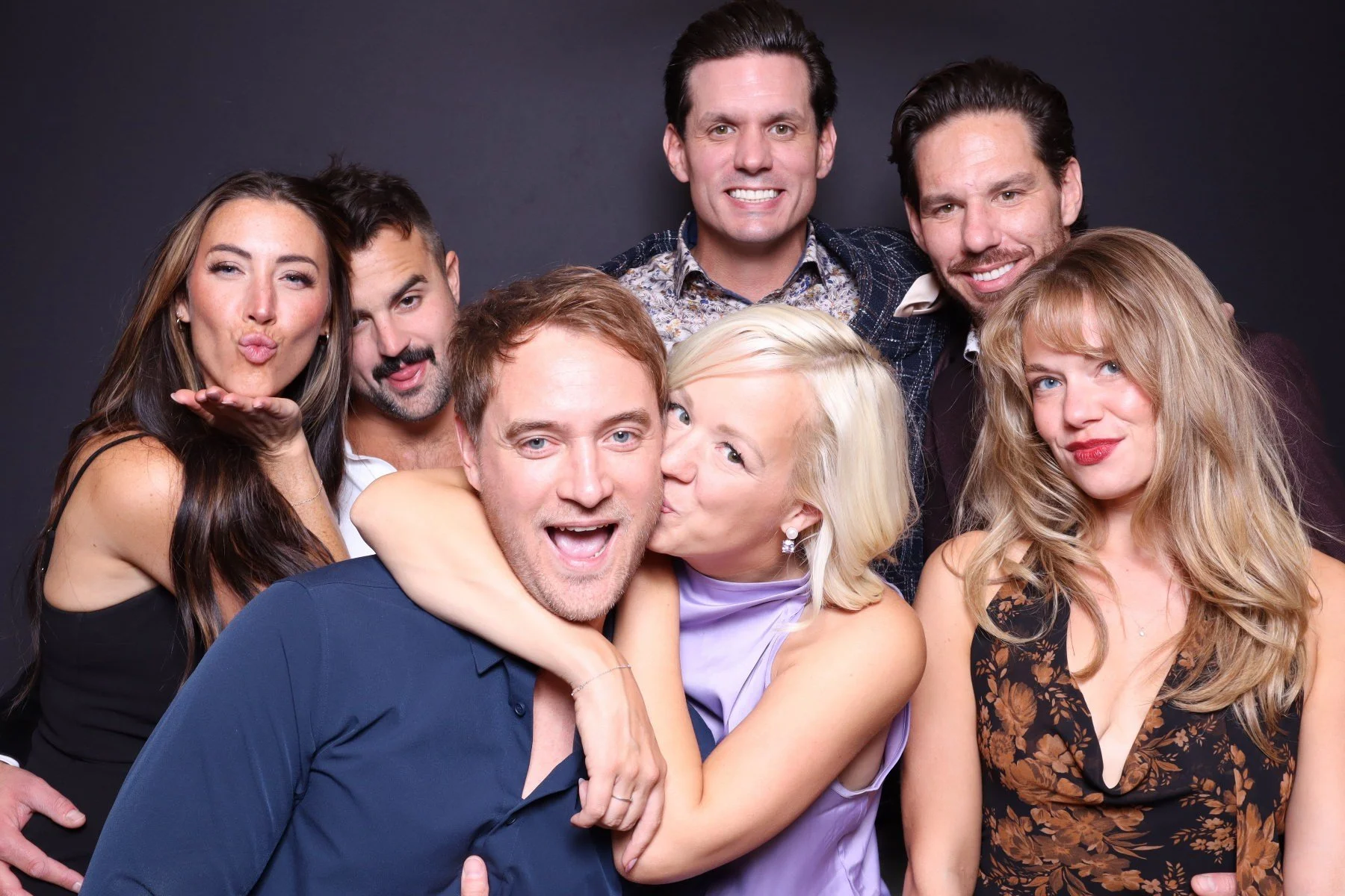 A group of seven diverse people, four women and three men, are smiling and posing for a photo against a dark background. One woman is giving a kiss and hug to a man in a blue shirt, while others are standing close together, some making playful expres