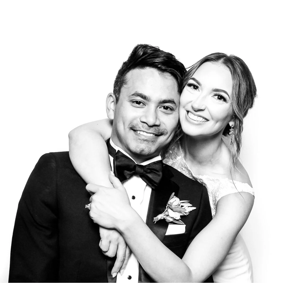 Black and white photo of a smiling couple, the man in a tuxedo and the woman in a wedding dress, embracing each other.