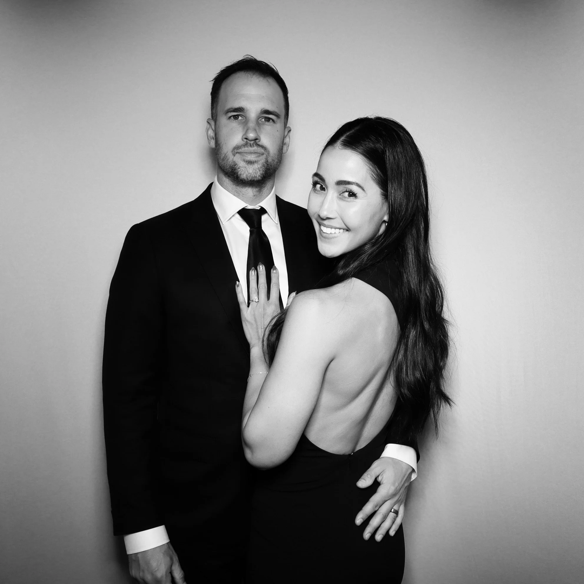 A black and white photo of a smiling woman with long dark hair and a man in a suit, standing against a plain wall. The woman shows her ring and is wearing a backless dress, and the man has his arm around her.