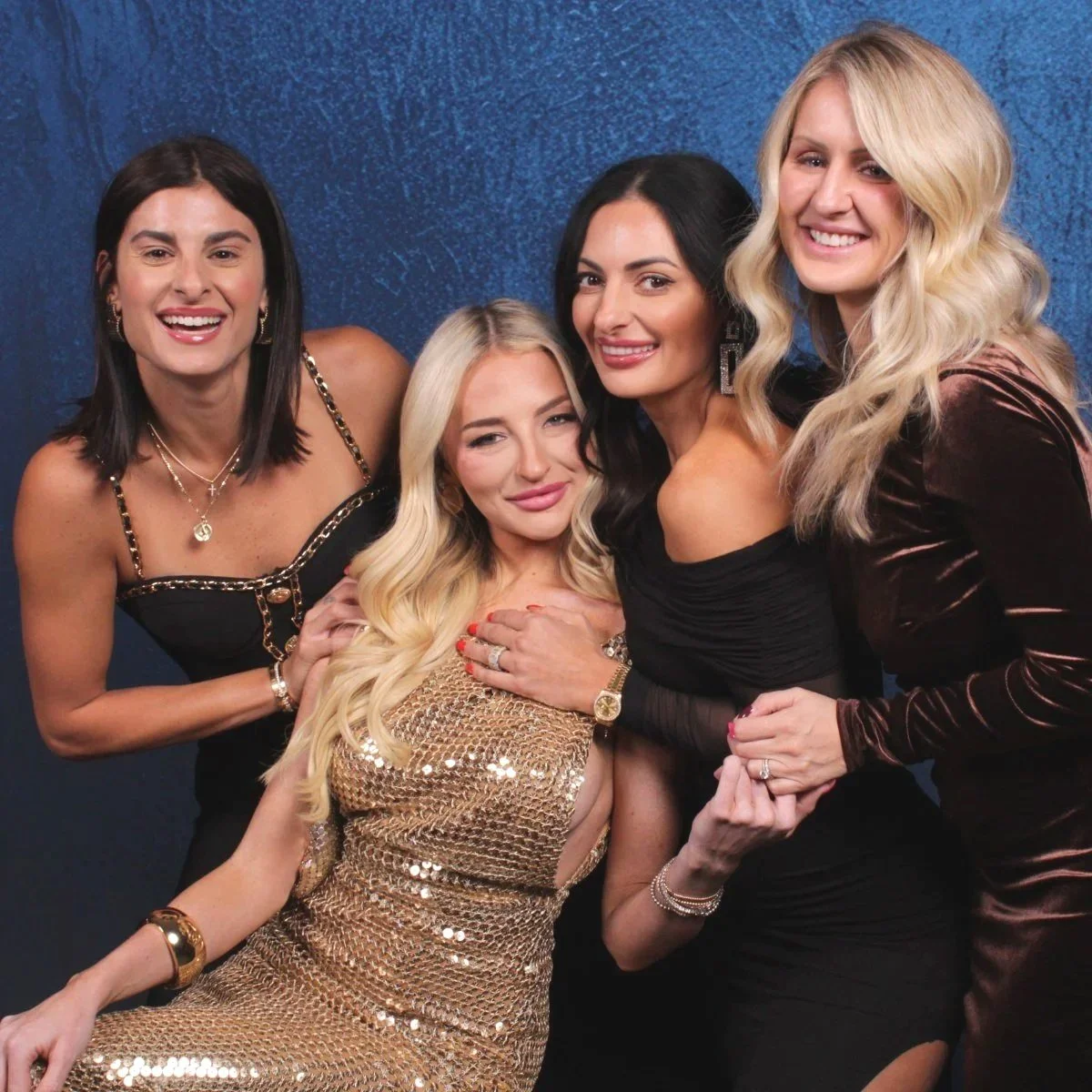 Five women dressed in glamorous outfits smiling and posing together against a dark blue background.