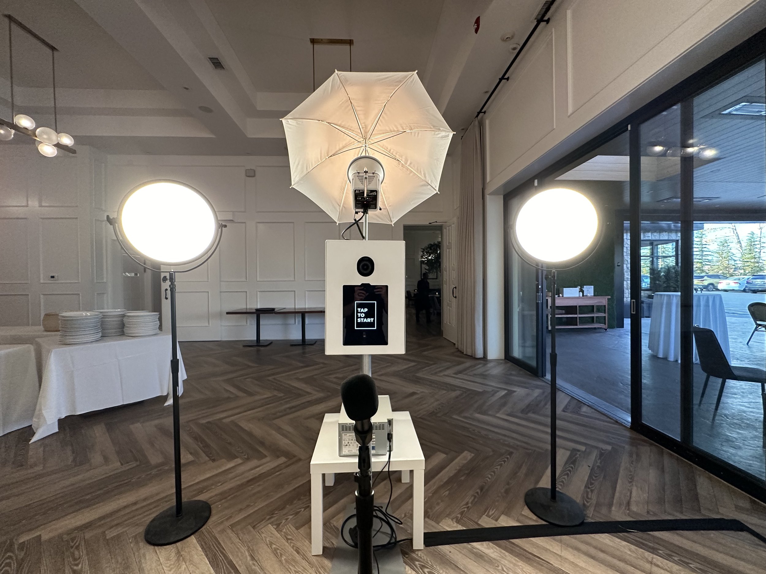Photo of photo booth setup with studio lighting, umbrella, camera, microphone, and screen in a spacious event room with wooden floors and sliding glass doors.