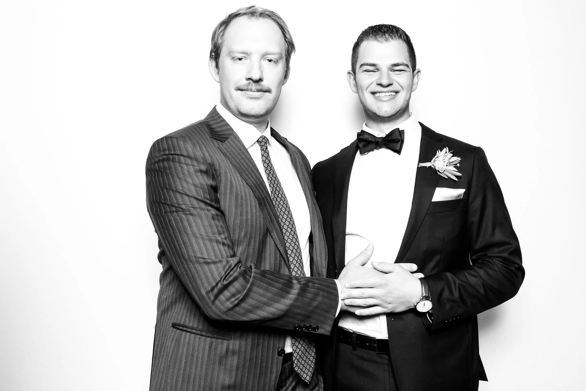 Two men dressed in formal suits, one in a pinstripe suit and the other in a tuxedo with a bow tie, smiling and holding hands against a plain background.