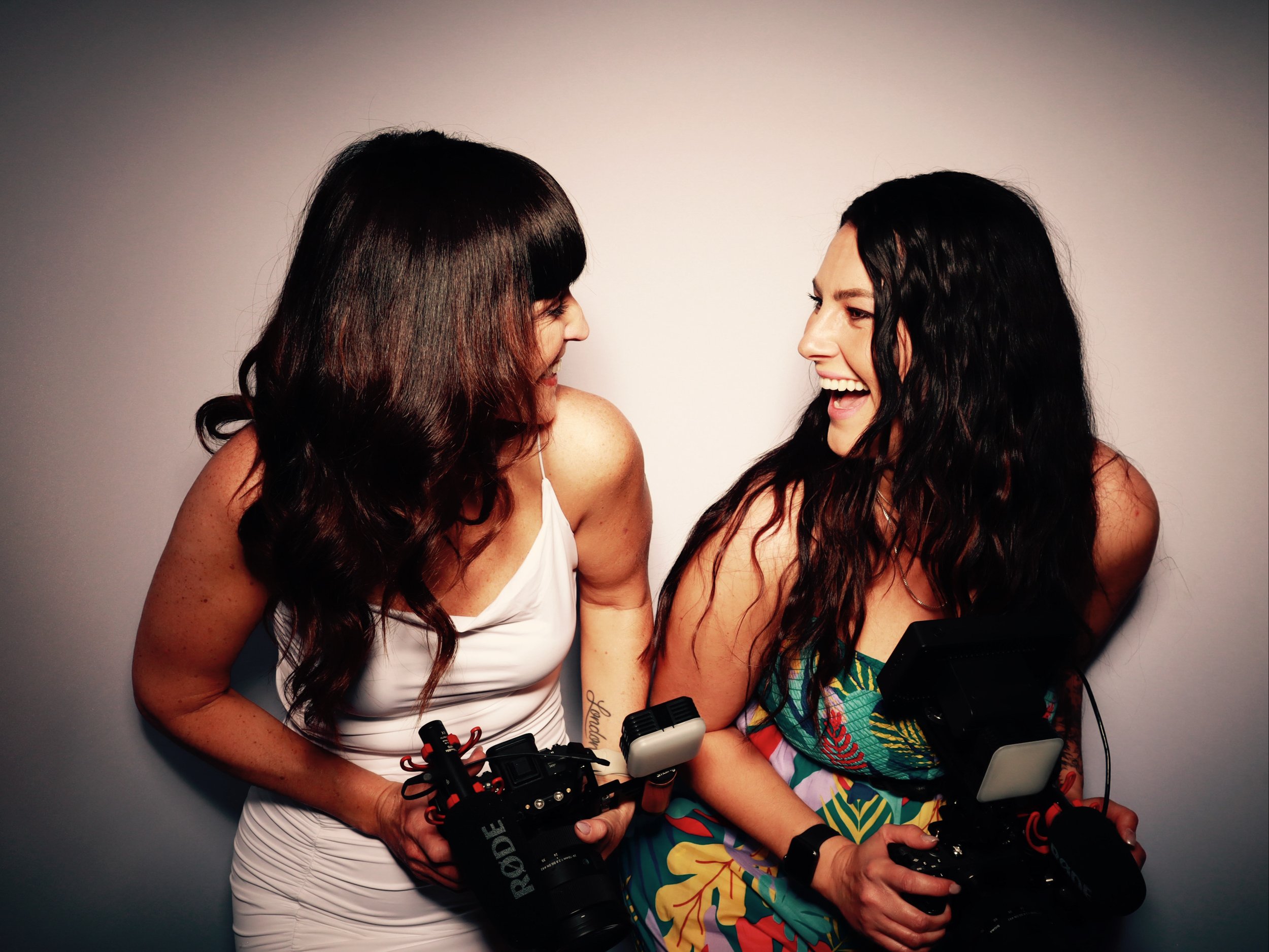 Two women with dark wavy hair laughing and holding cameras against a plain wall.