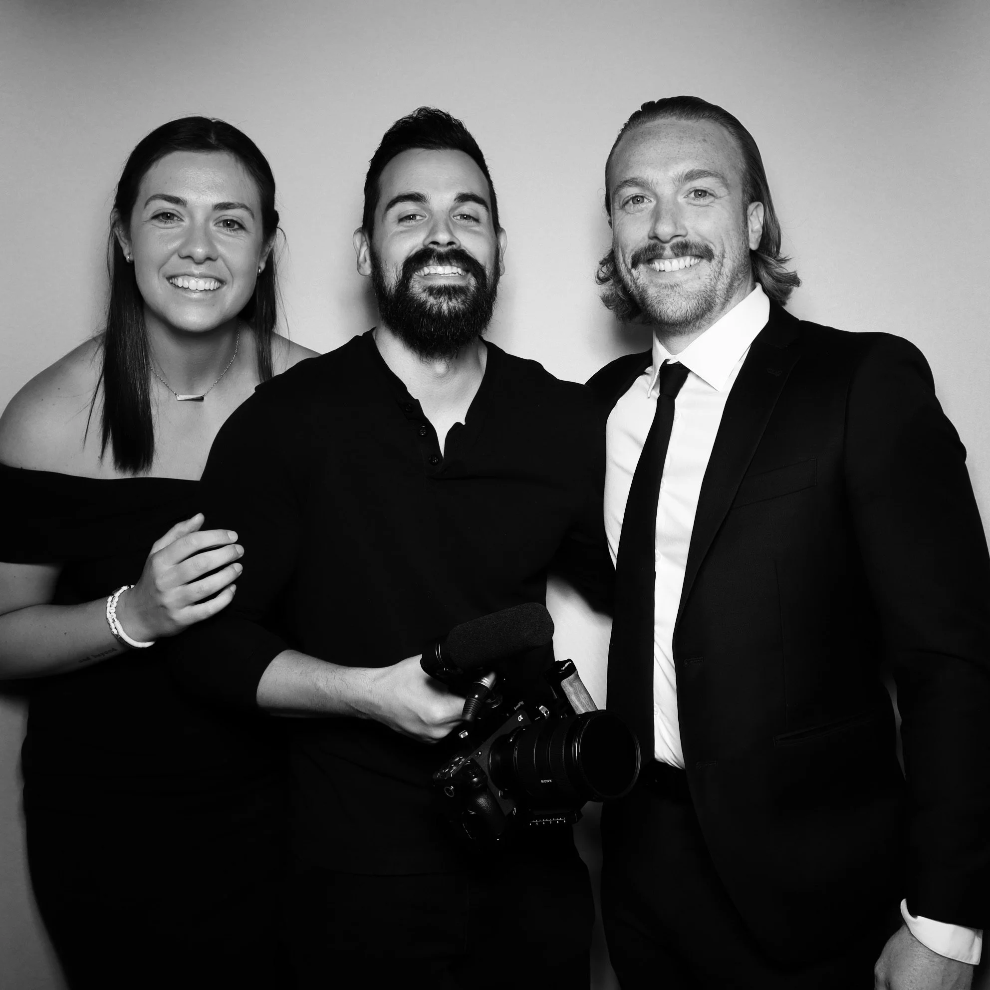 Black and white photo of three smiling people in formal attire standing close together. The woman on the left has shoulder-length dark hair and wears an off-the-shoulder dress. The man in the middle has a beard, short hair, and holds a camera with a 