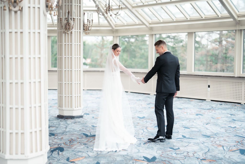 A bride and groom holding hands in a bright, glass-enclosed room with patterned carpet.