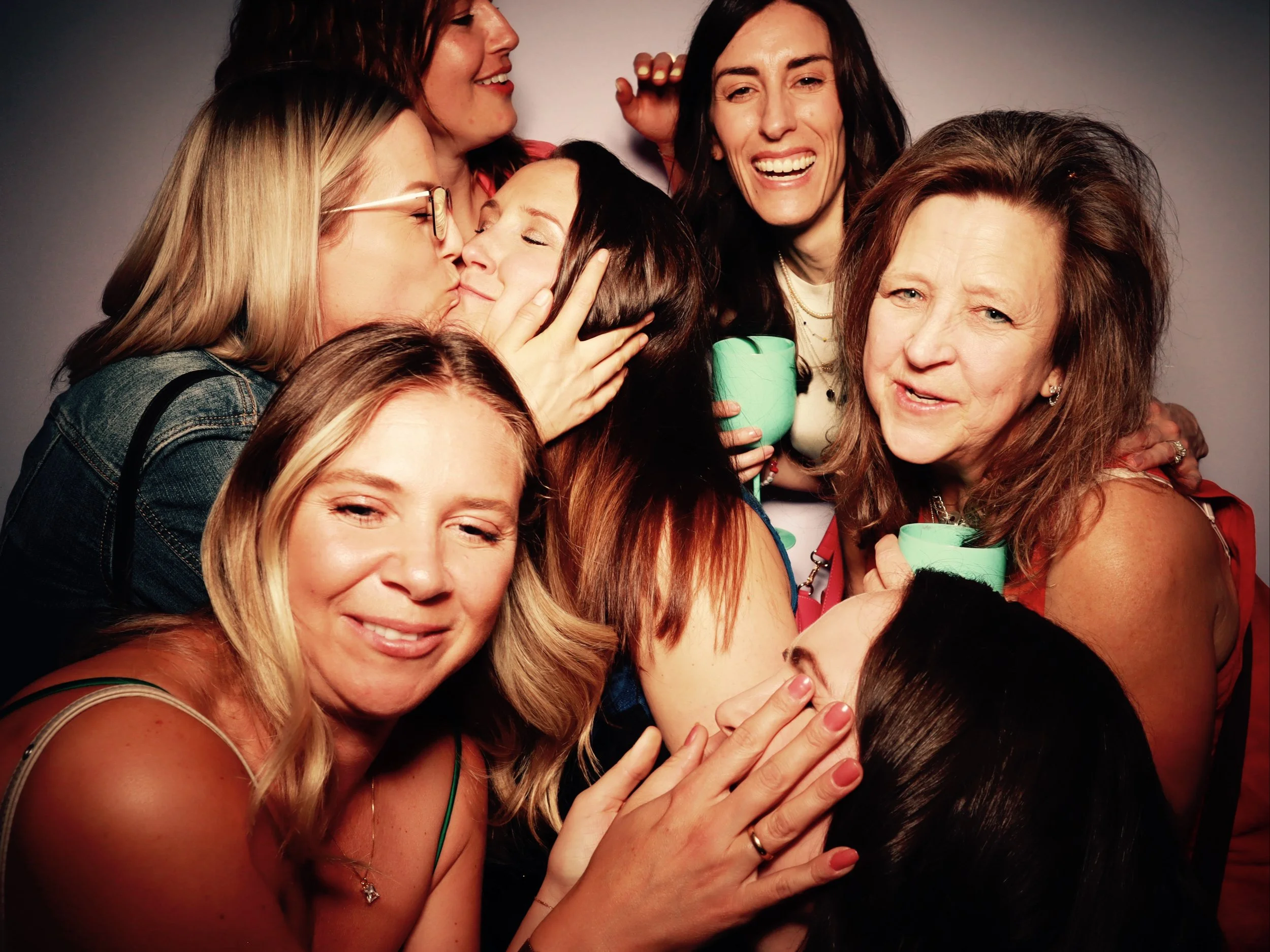 A group of eight women engaging in affectionate and joyful interactions, including kissing, hugging, and smiling, against a plain background.