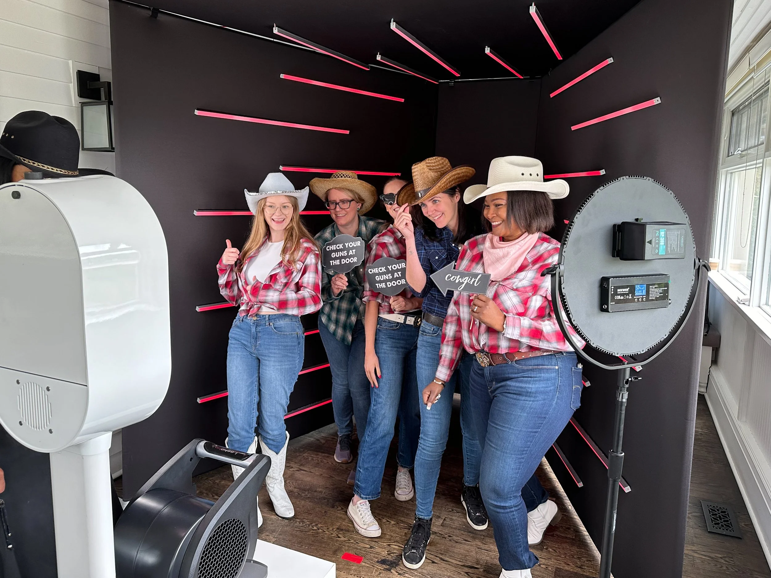 Five women dressed in plaid shirts and jeans wearing cowboy hats, posing for a photo with some holding signs that say 'Check your guns at the door' and 'Cowgirl'. They are standing in front of a black wall with pink light bars and a photo booth setup