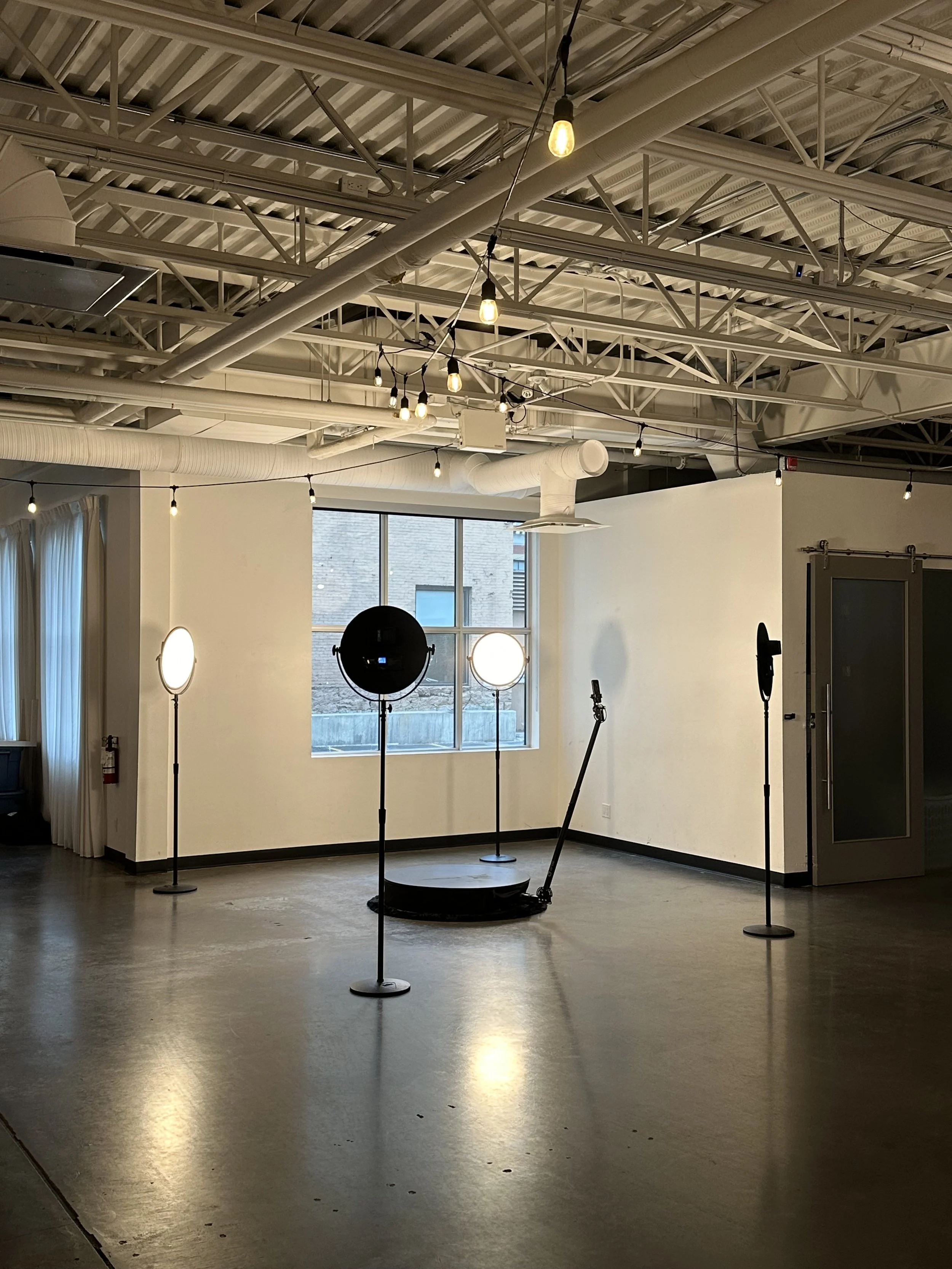 Empty indoor space with a 360 video booth professional lighting and camera equipment, including three ring lights on stands, a black stage platform, and a microphone stand, with a window showing a brick building outside.