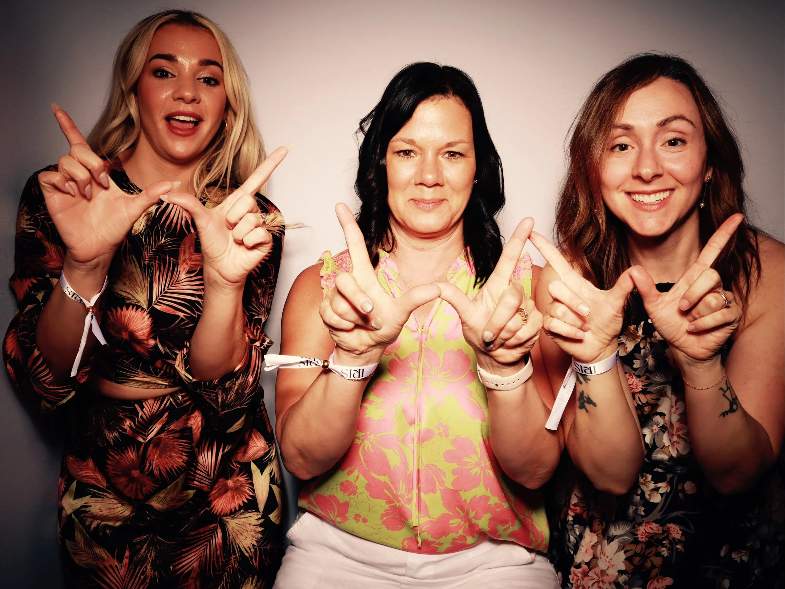 Three women standing side by side making hand signs, smiling, and wearing wristbands against a plain wall.