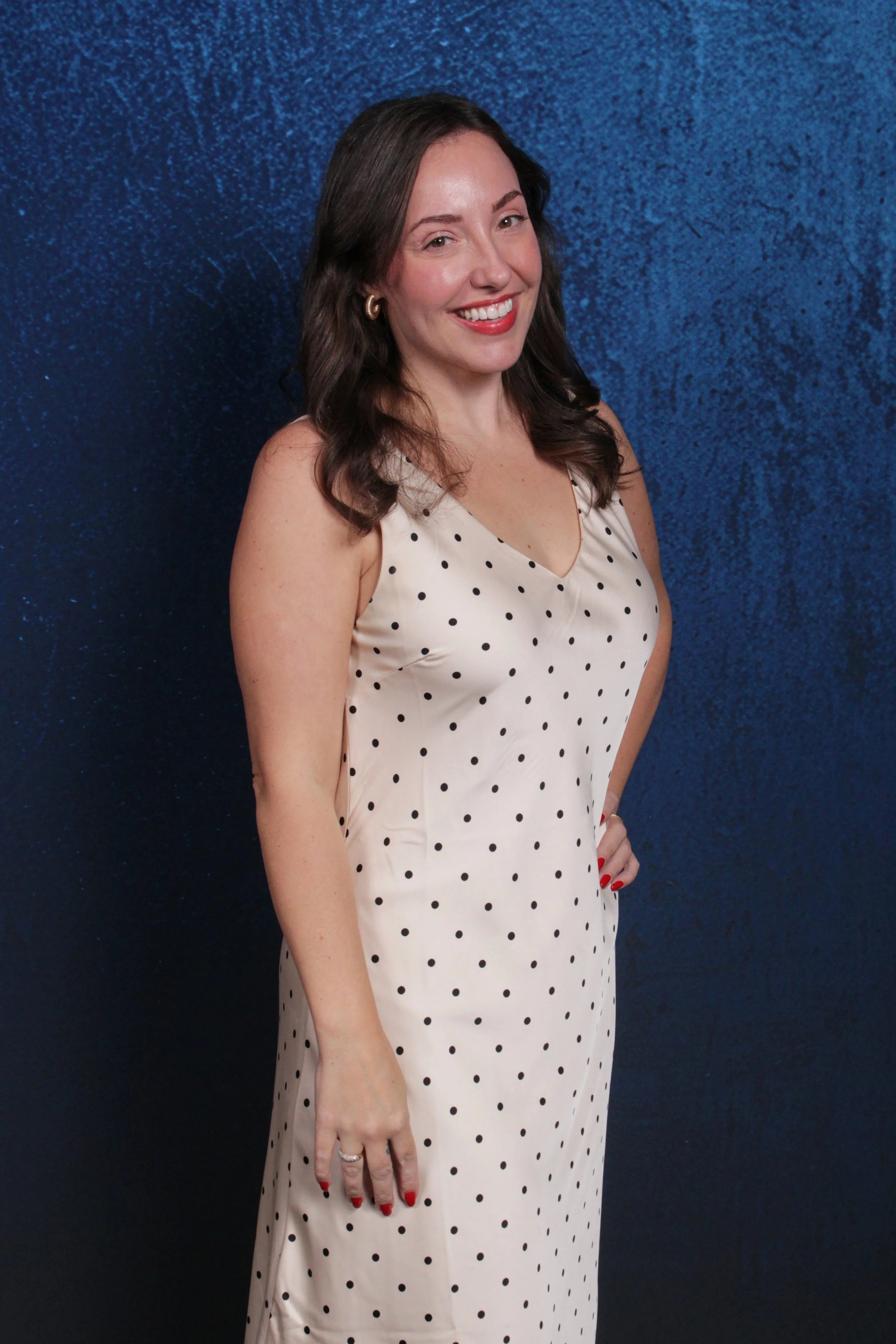 A woman with shoulder-length brown hair, wearing a beige dress with black polka dots, standing against a textured blue background.