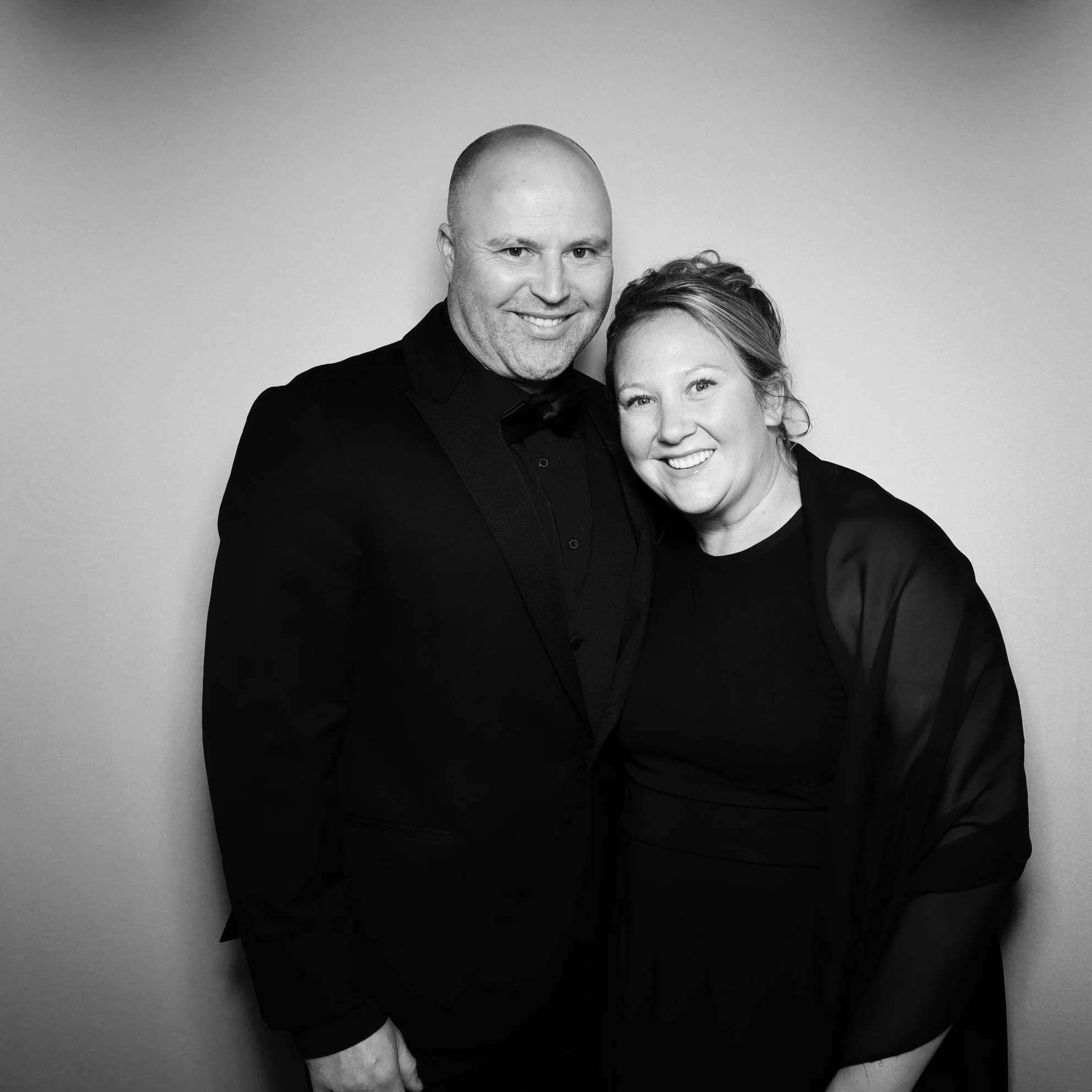 A black and white photo of a man wearing a tuxedo and a woman in a black dress, smiling and standing close together in front of a plain background.