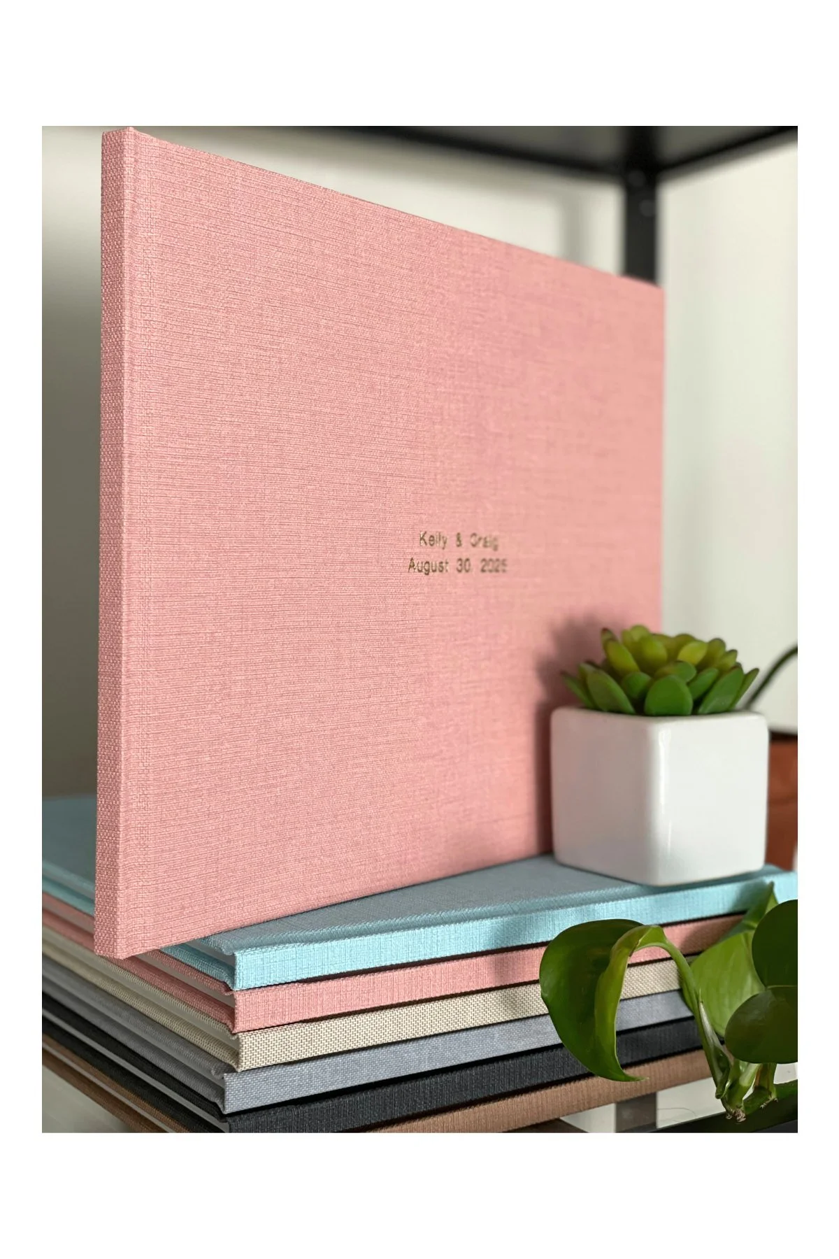 A pink photo album with engraved text and a small potted succulent plant, positioned on a stack of pastel-colored books on a table.