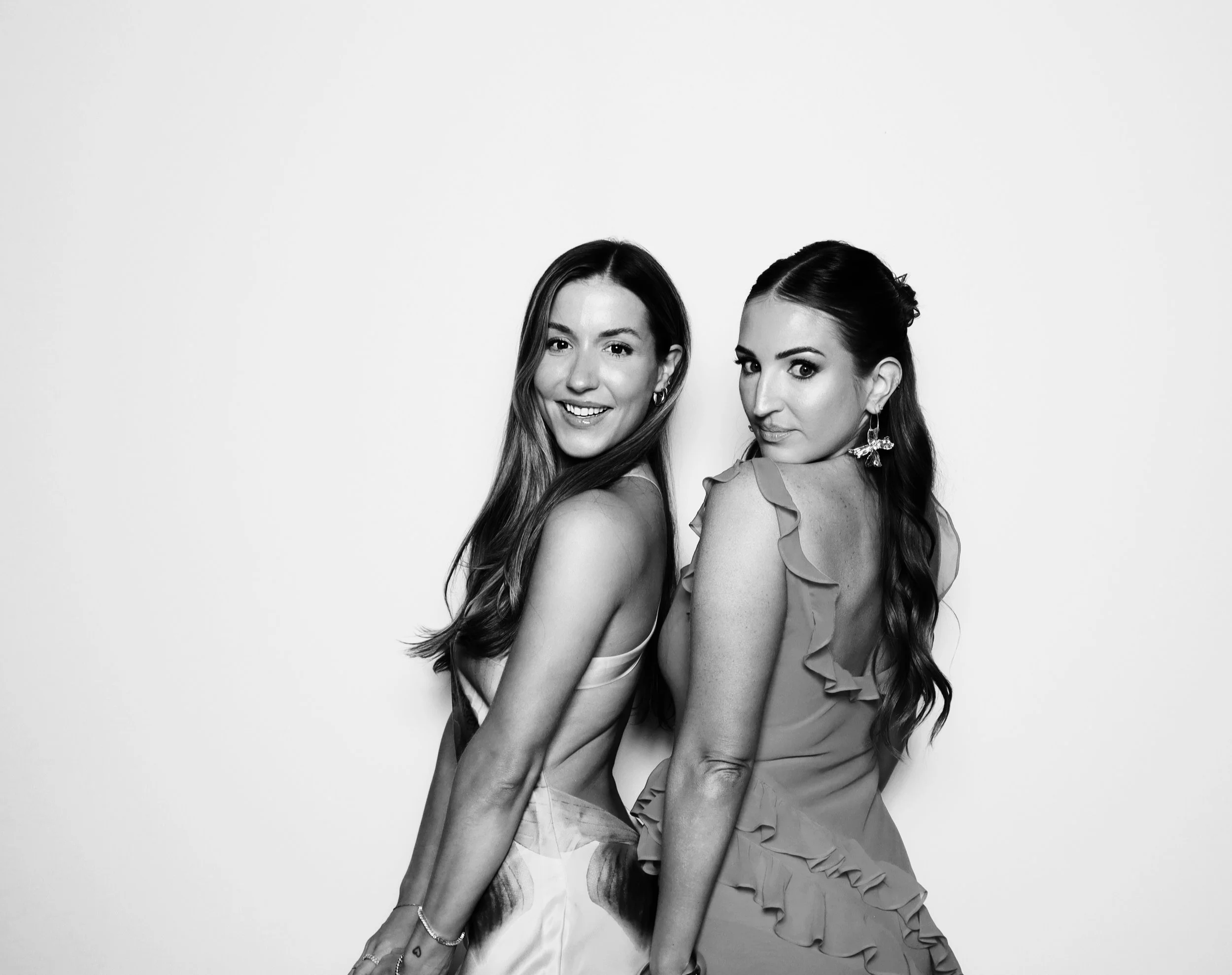 Two women posing against a plain background, smiling and looking at the camera, in black and white. One woman wears a light-colored dress with ruffles on the sleeves, and the other woman wears a sleeveless top or dress, both with long hair.