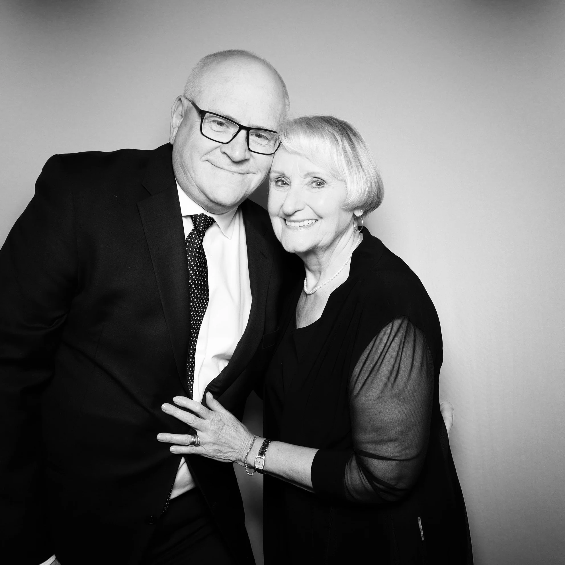 A black and white photo of an older man and woman smiling and posing together, dressed in formal attire, against a plain background.