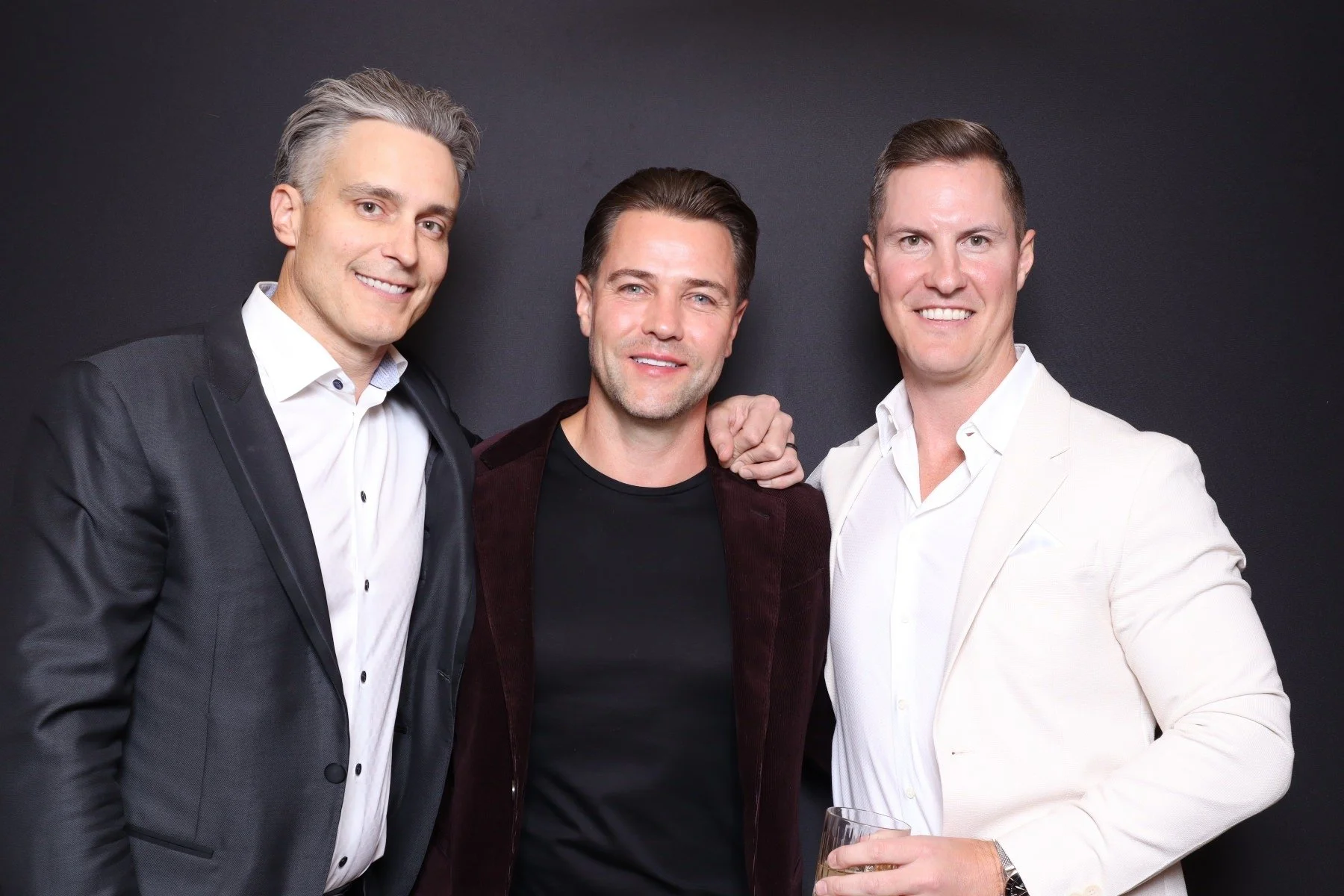 Three men in formal attire standing close together against a black background, smiling at the camera. The man on the left is wearing a black suit and white shirt. The man in the middle is wearing a dark jacket and black shirt. The man on the right is