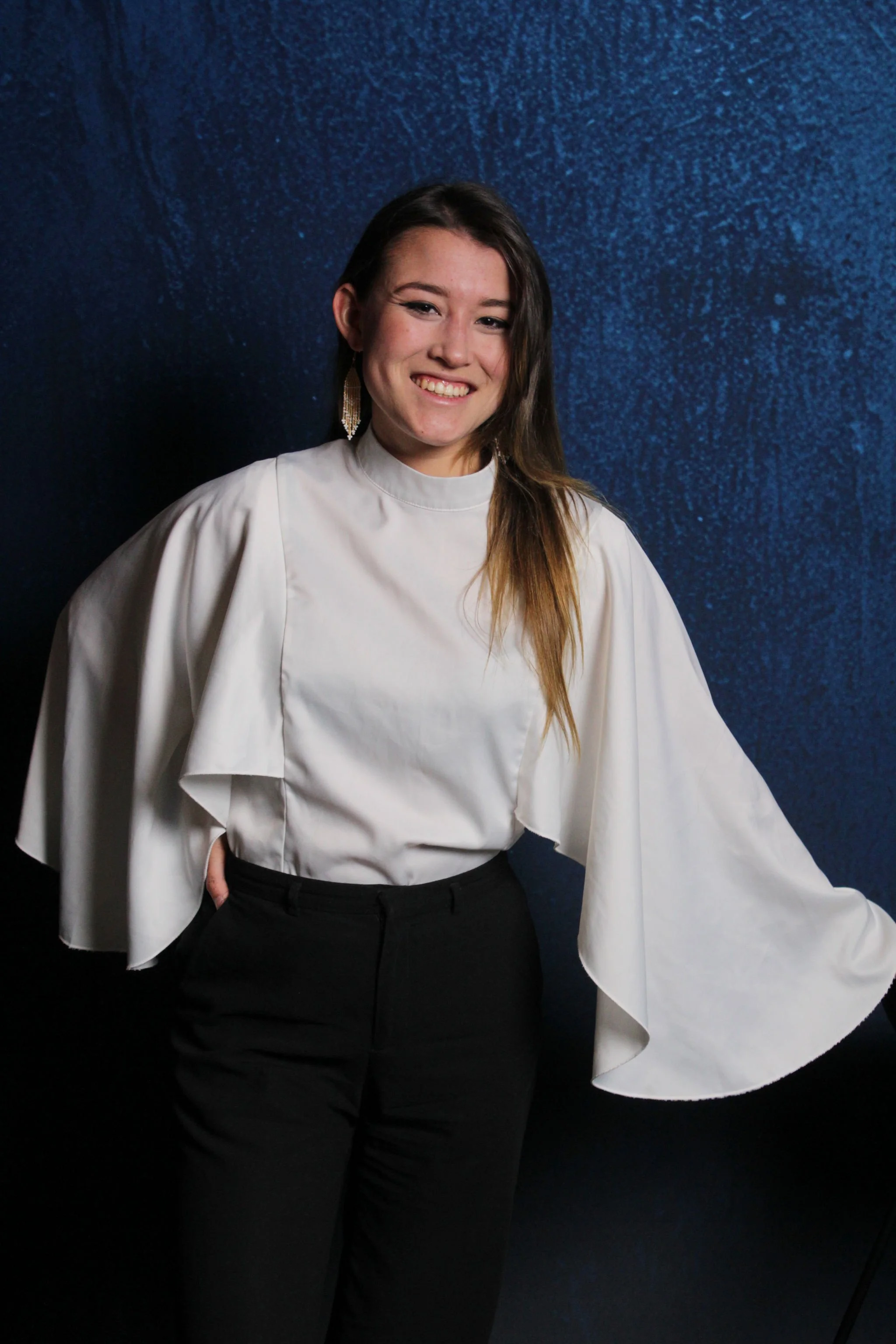 A young woman with long brown hair, wearing a white top with dramatic, wide sleeves and black pants, standing against a dark textured blue background, smiling.
