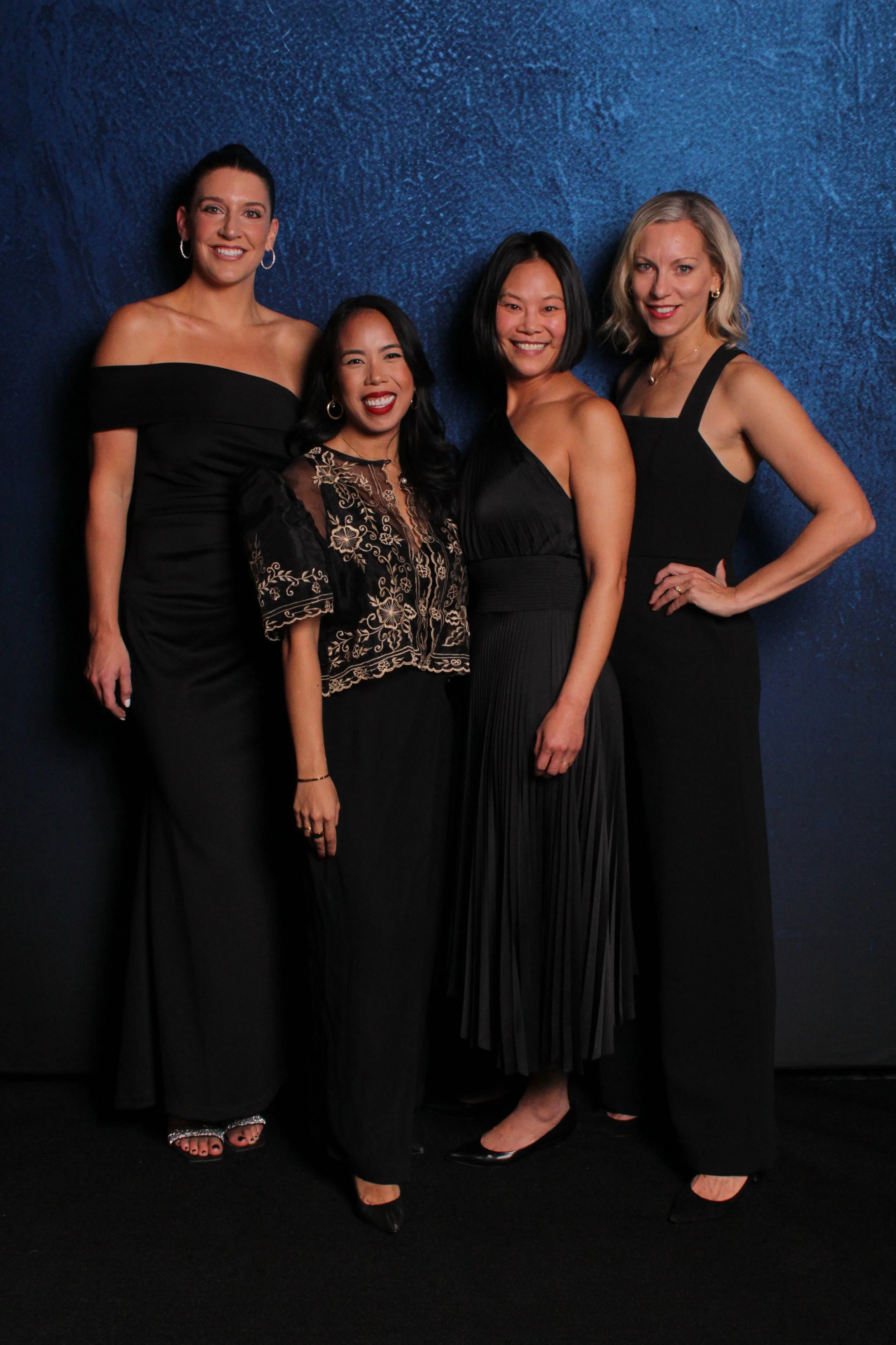 Group of four women in black evening gowns posing against a blue textured wall