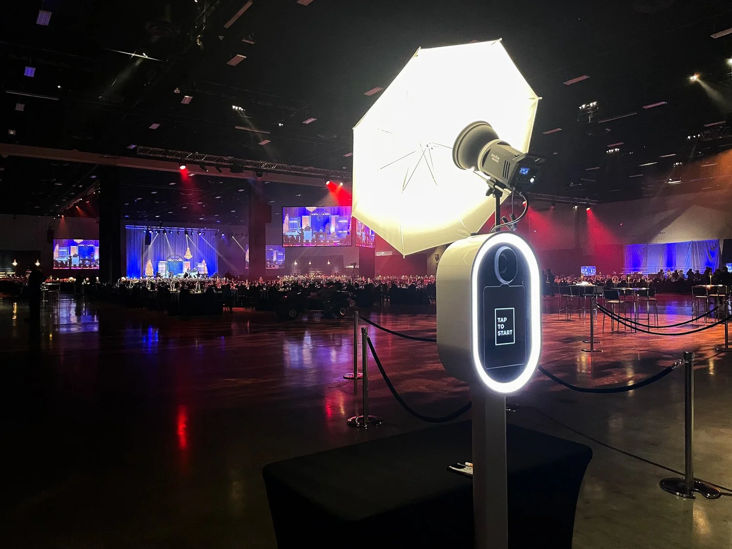 A photo of a large banquet hall with a stage and many round tables. In the foreground, there is a selfie station with a ring light, a camera, and a white umbrella softbox for photography. The hall is dimly lit with blue, red, and purple stage lightin