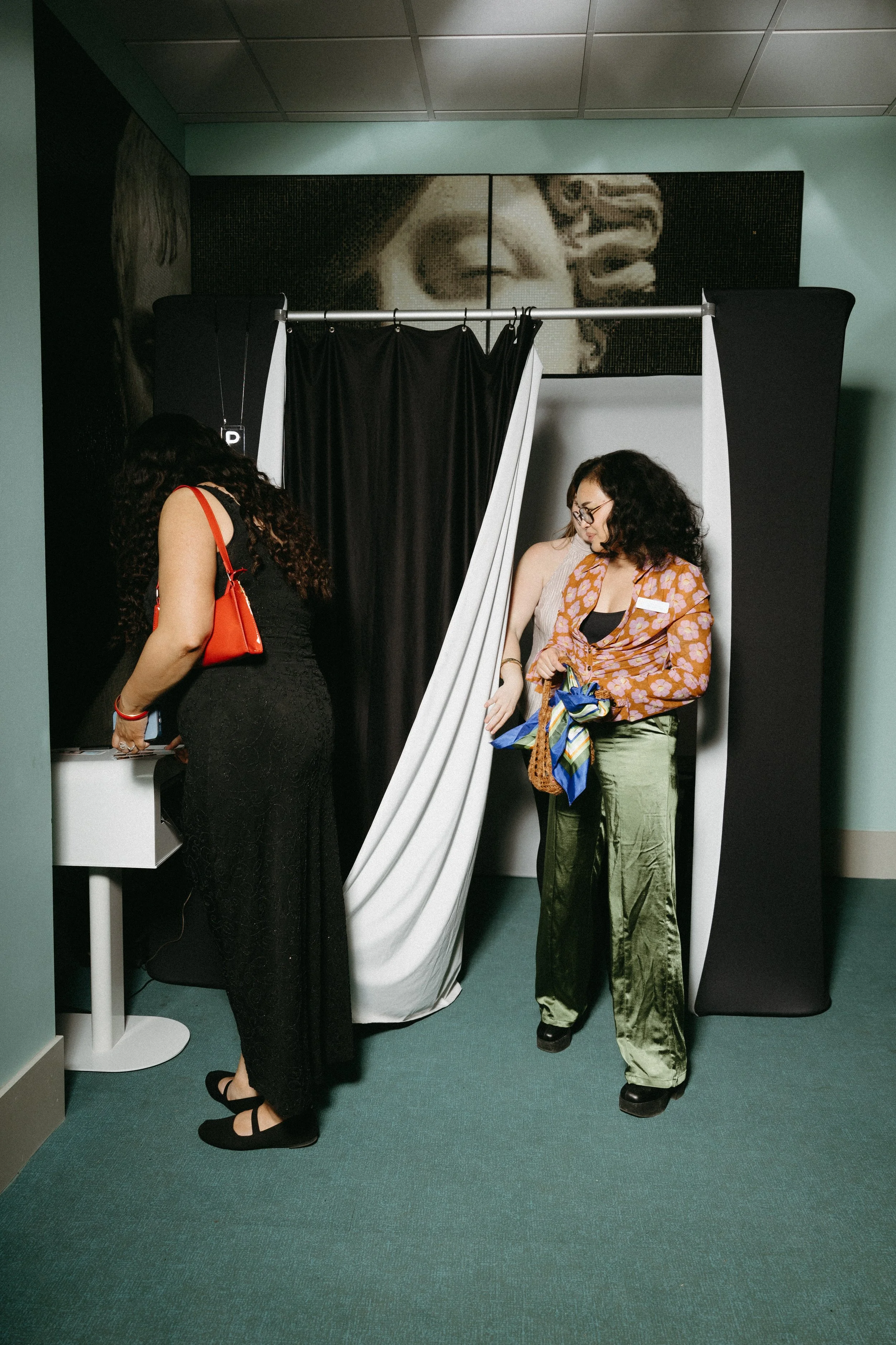 Two women are participating in a photo booth session, with one woman adjusting her purse and the other looking down at her purse or phone, inside a room with a backdrop featuring a partial artwork or mosaic of a face.