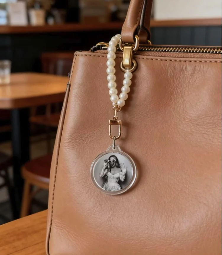 A tan leather handbag with a gold zipper and a pearl strap attached. The pearl strap has a circular keychain with a black and white photo of a woman wearing sunglasses and a lace dress, holding a puppy. The background shows wooden tables and chairs i