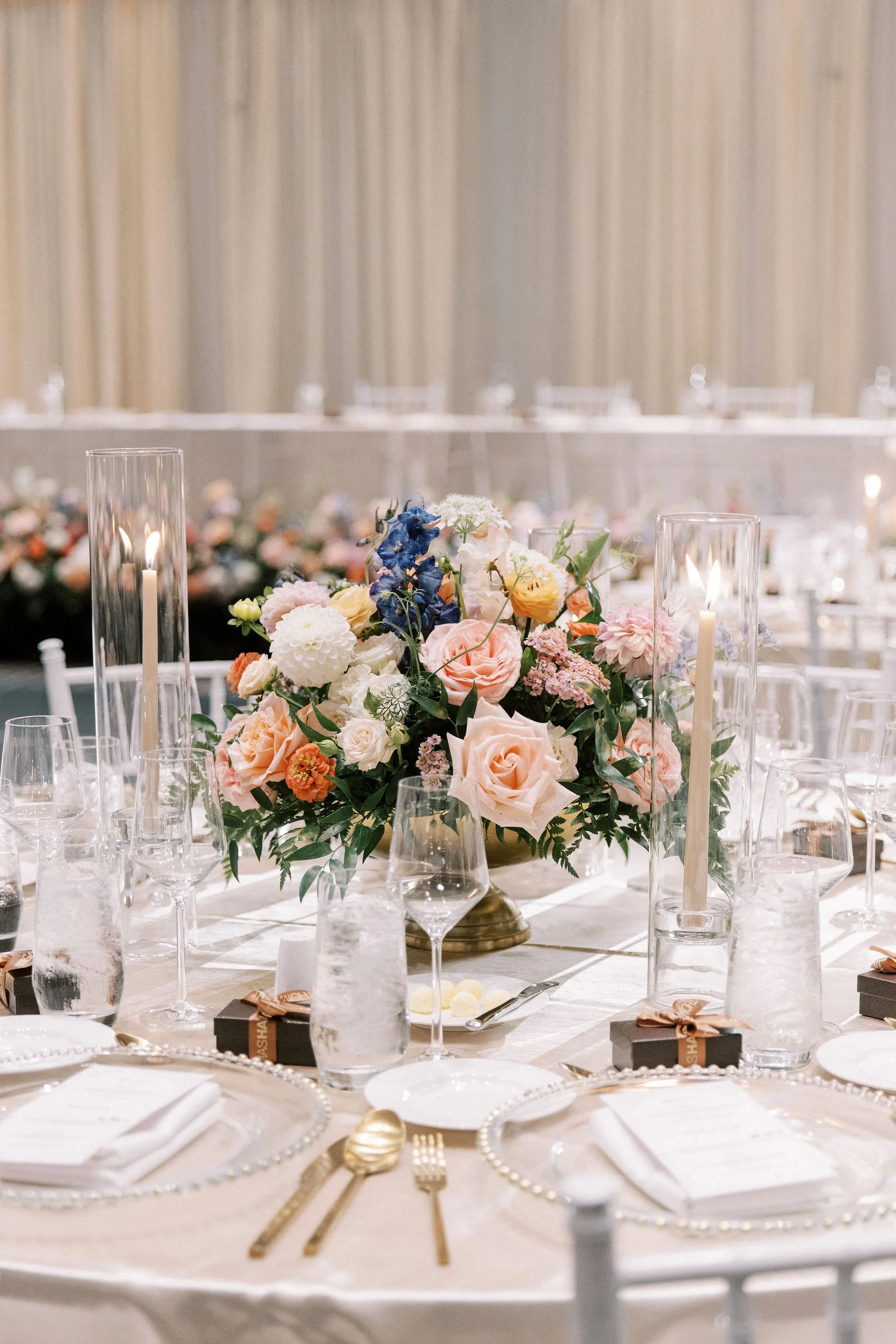 Elegant banquet table decorated with a large bouquet of pink, white, and blue flowers, surrounded by tall glass candleholders with candles, glassware, and wrapped party favors.