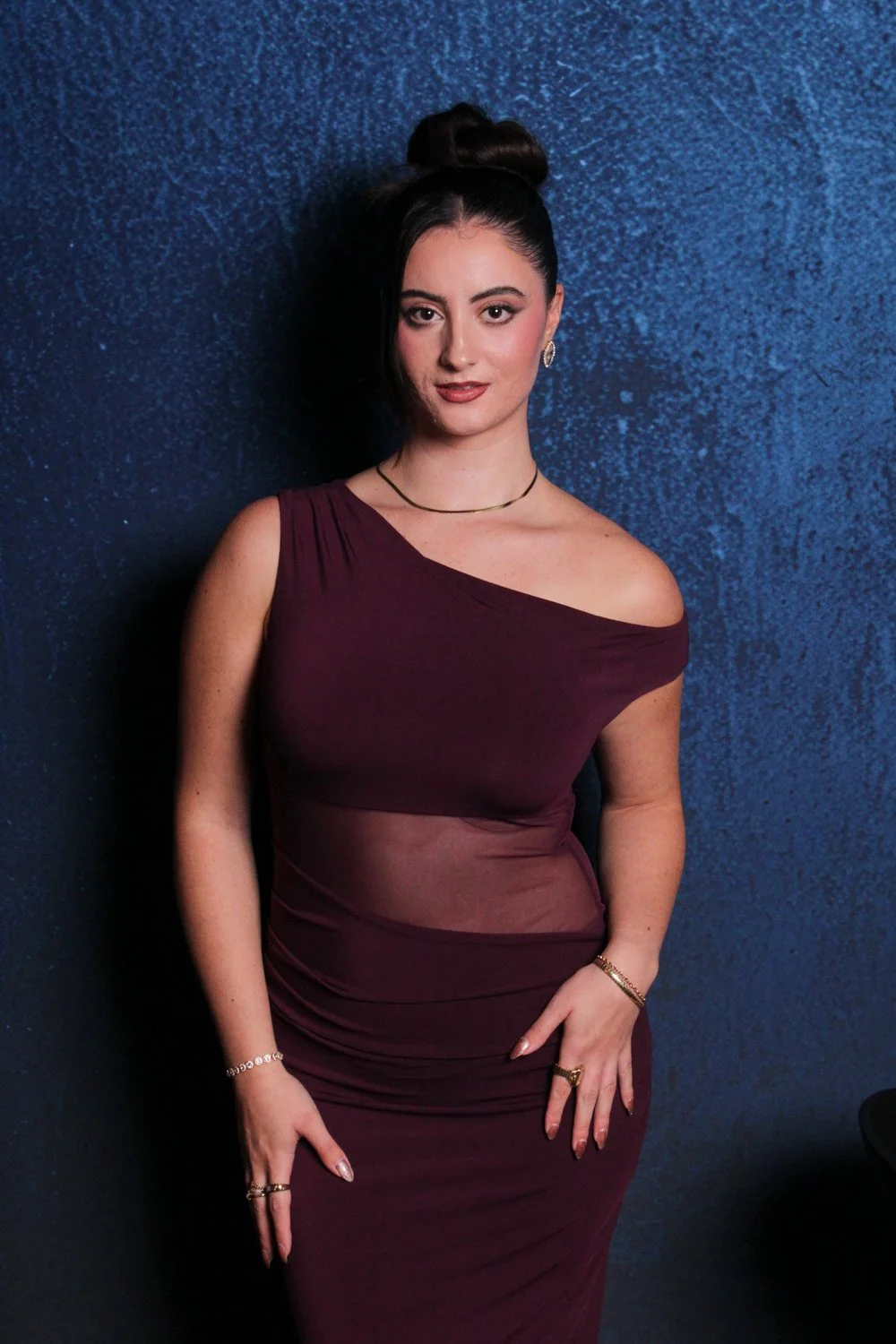 A woman standing against a dark blue, textured wall, wearing a burgundy one-shoulder dress with sheer section at the waist, her hair styled in a high bun with a long side section, accessorized with earrings, a necklace, and rings.