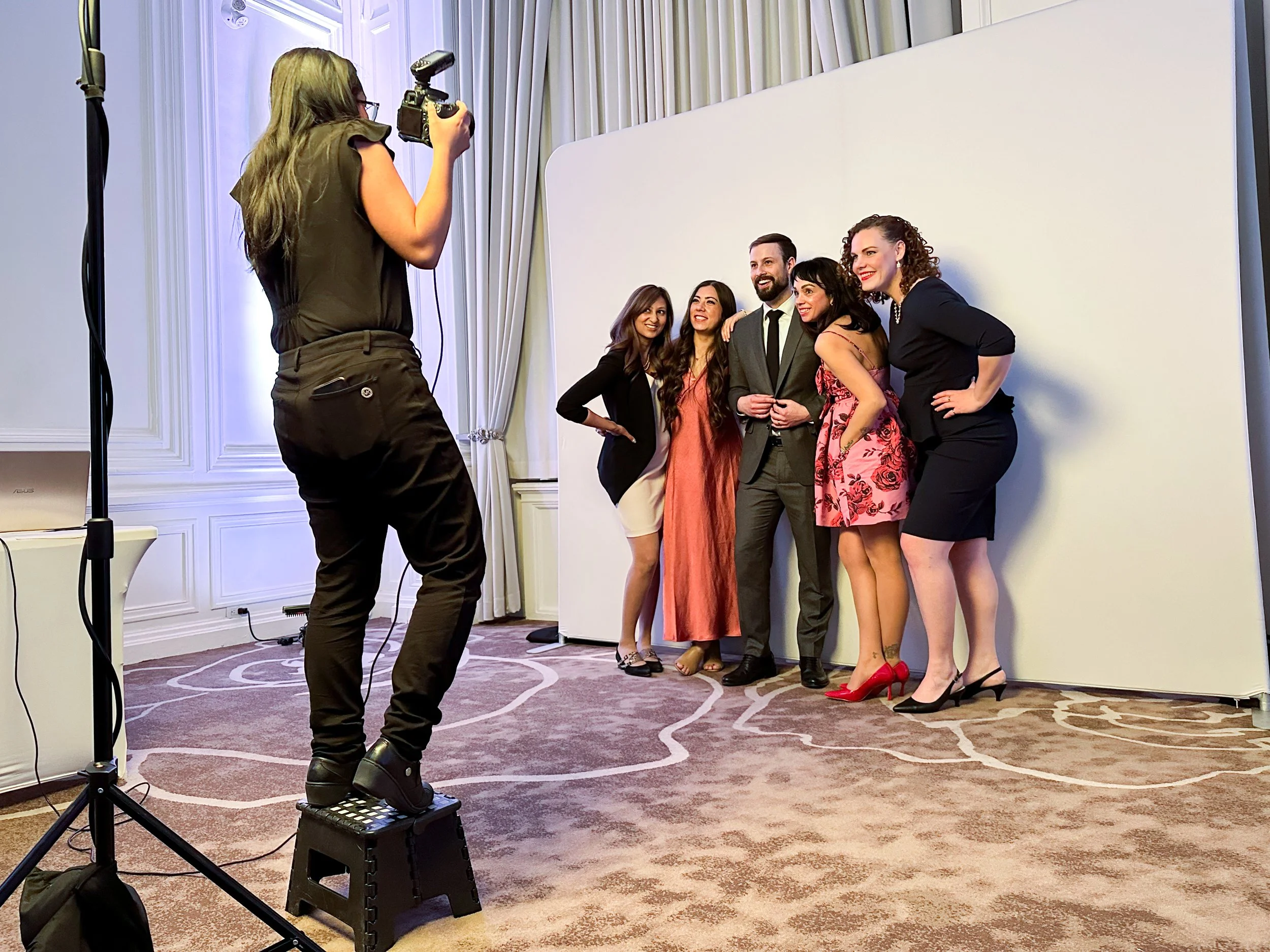 Group of five people posing for a photo in front of a white backdrop, with a photographer taking their picture, inside a well-lit room with curtains and elegant wall paneling.