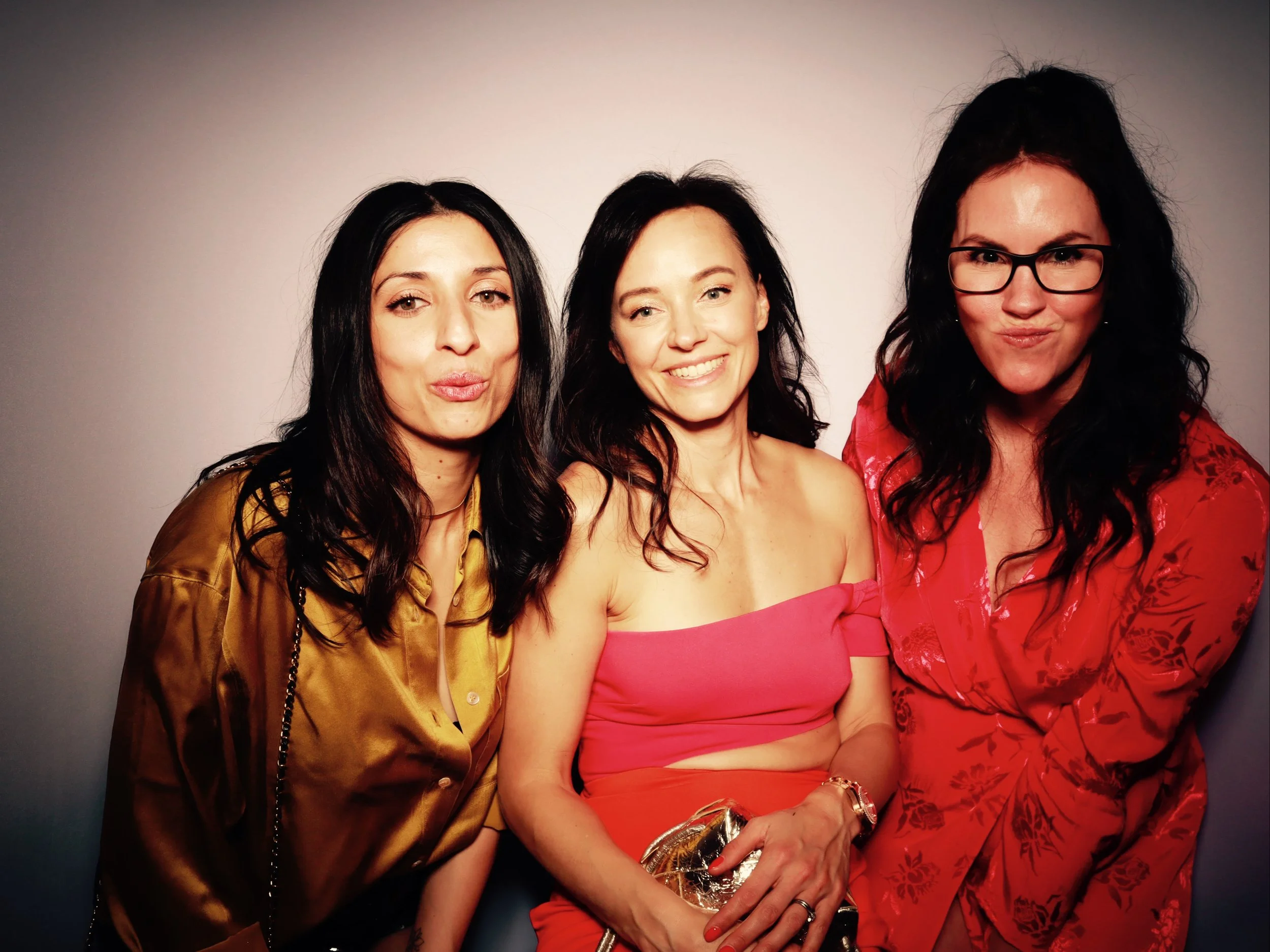 Three women with dark hair posing together against a gray background, dressed stylishly, smiling and making playful expressions.