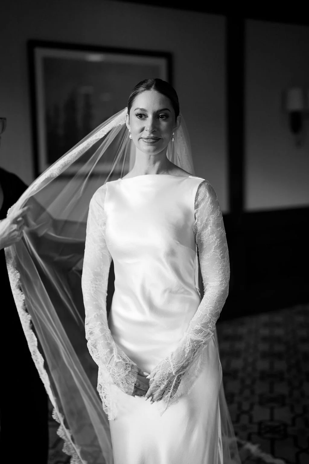A bride in a satin wedding dress with lace sleeves stands indoors, adjusting her veil, smiling gently.