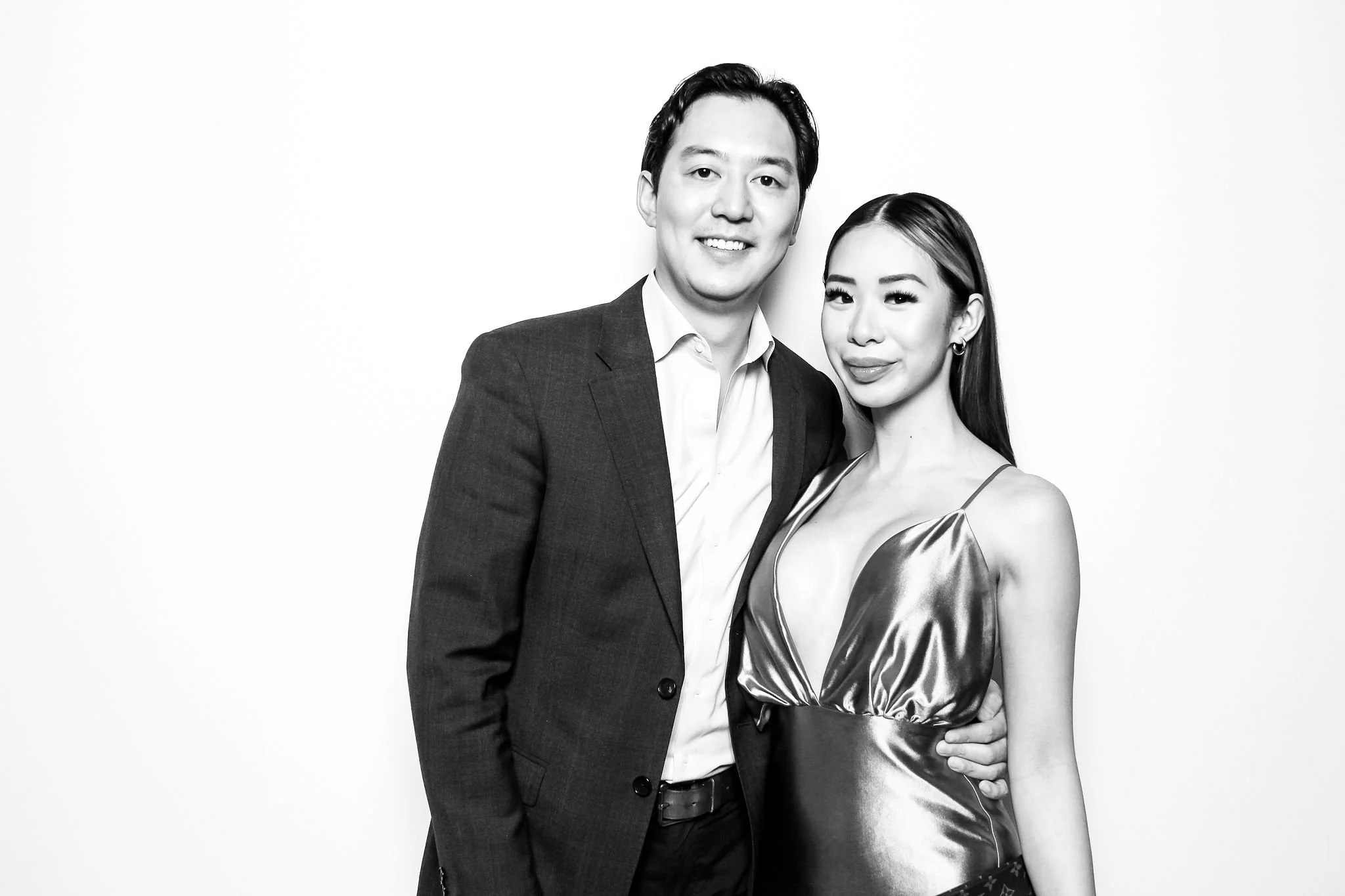 A black and white photo of a smiling man in a suit and a woman in a shiny, deep V-neck dress standing close together against a plain white background.