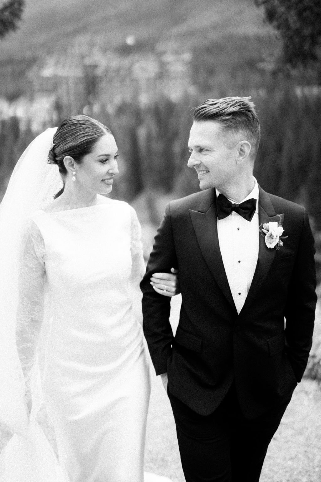 A black and white photo of a bride and groom on their wedding day. The bride is wearing a white dress and veil, and the groom is dressed in a tuxedo with a bow tie and boutonniere. They are smiling at each other outdoors.