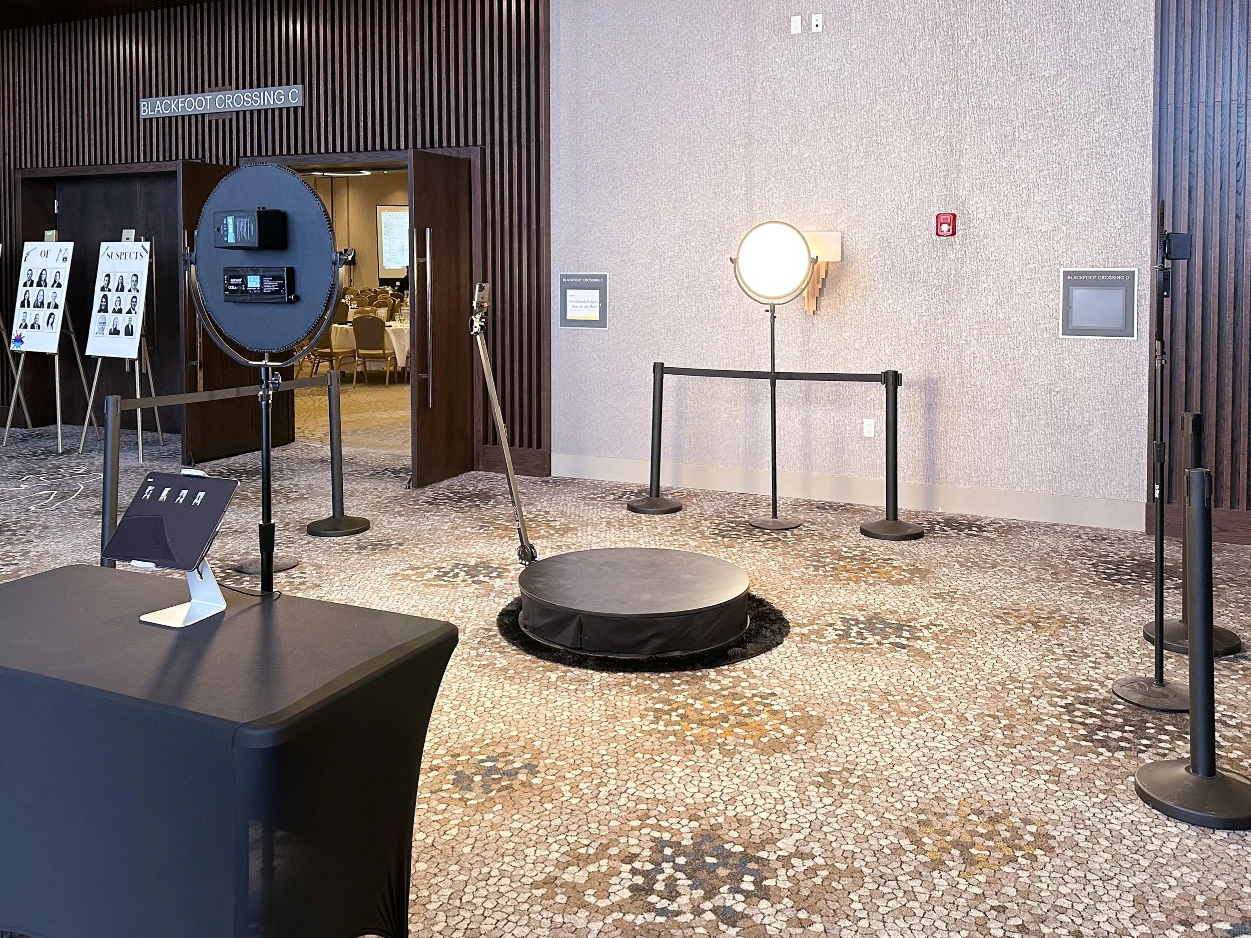 Photo of a photo booth station set up in a conference space with lighting equipment and a circular platform for photos, near a hallway leading to a room filled with round tables and chairs.