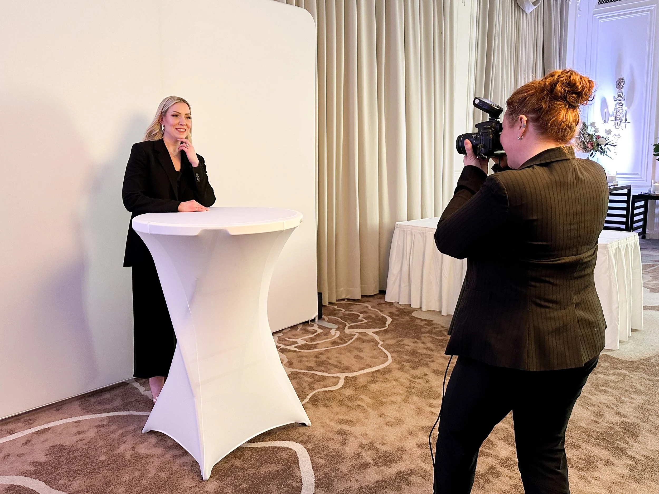 A woman is being photographed at a high-top table in a room with cream-colored curtains and patterned carpet.