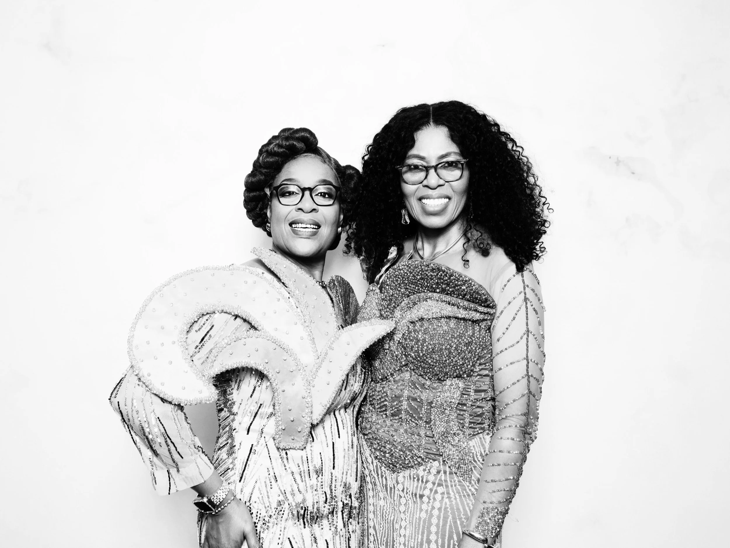 Two women dressed in elaborate, patterned outfits posing together against a plain background, smiling at the camera.