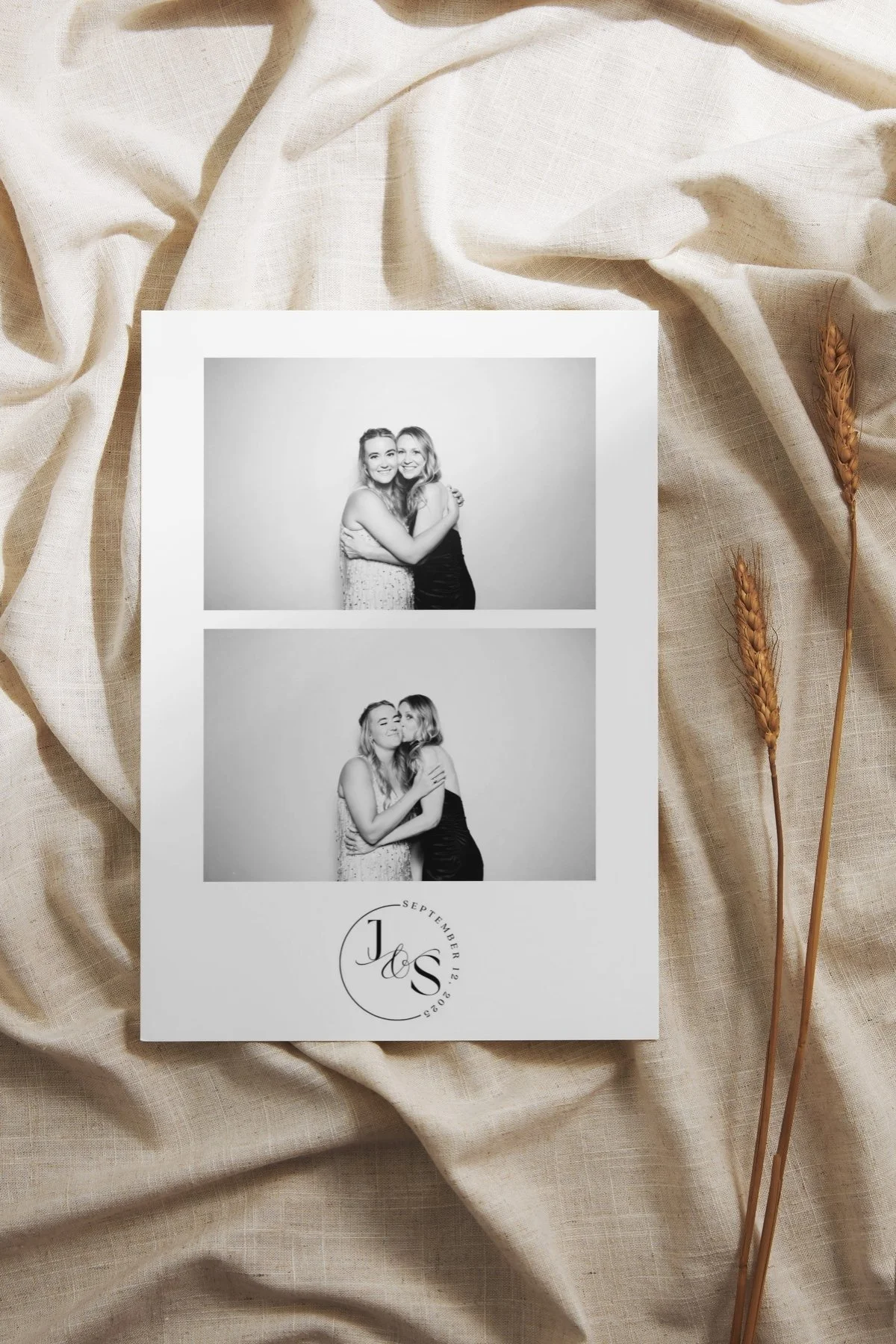 A photo strip with two black and white images of two women hugging. The photo strip has a monogram at the bottom with initials 'J & S' and the date September 12, 2023, against a backdrop of beige fabric with dry wheat stalks.