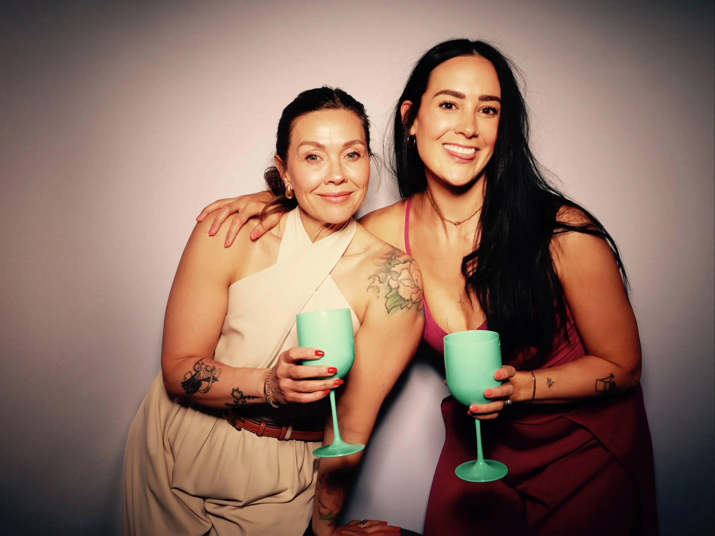 Two women smiling and holding turquoise wine glasses, standing close together against a plain background.