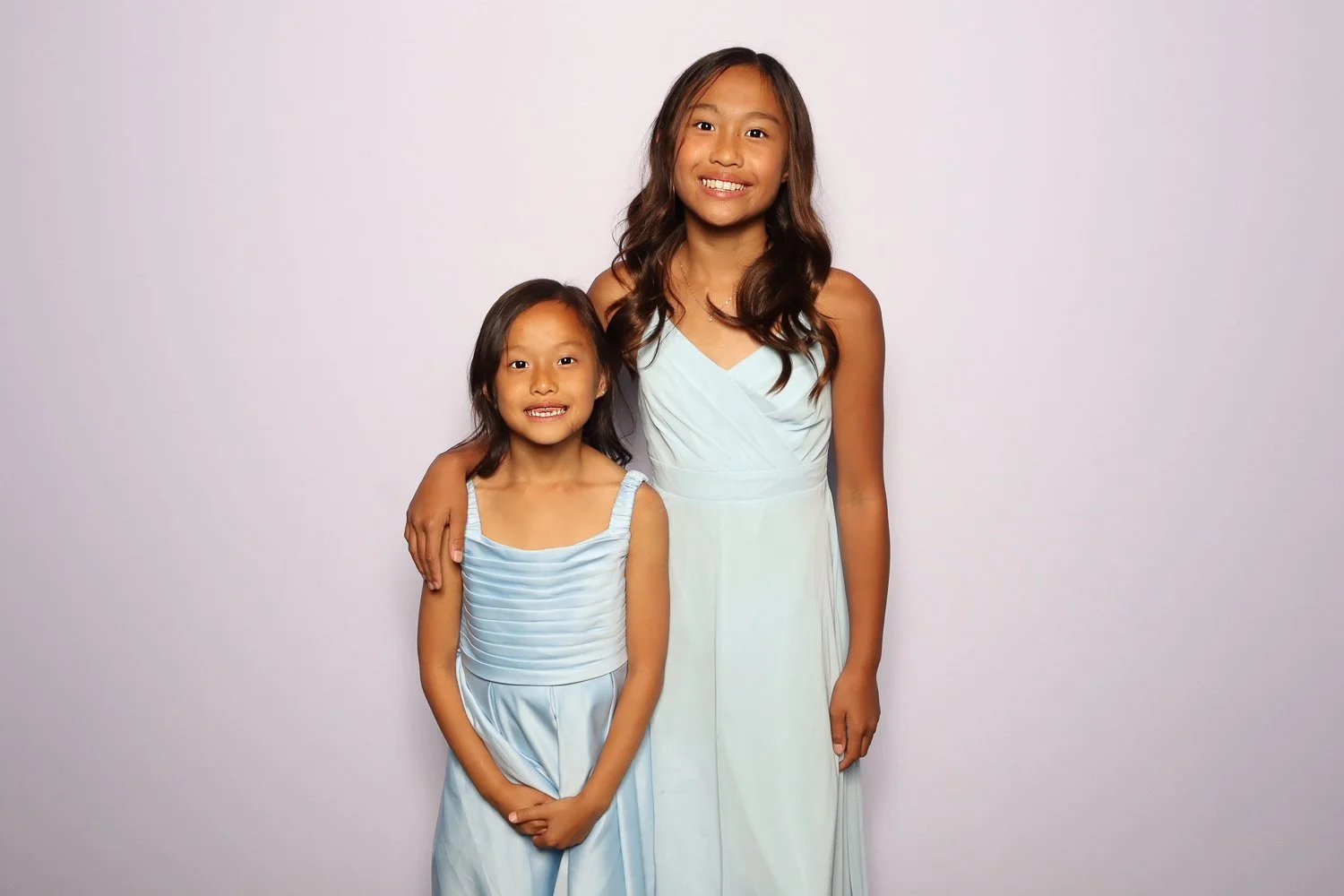 Two young girls dressed in light blue dresses, standing close together, with the older girl’s arm around the younger girl’s shoulder, smiling in front of a plain light background.