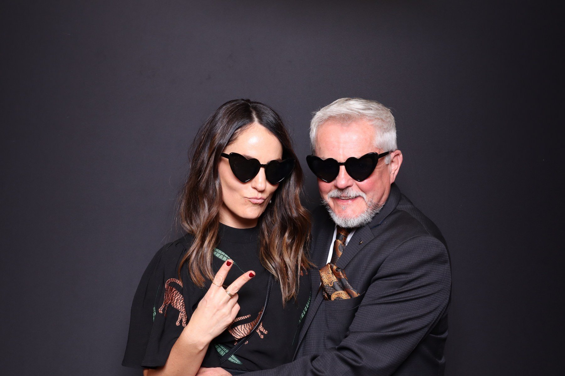 A woman and an older man wearing black sunglasses shaped like hearts, posing together against a dark background. The woman is making a peace sign and has long brown hair, while the man has short gray hair and a beard.
