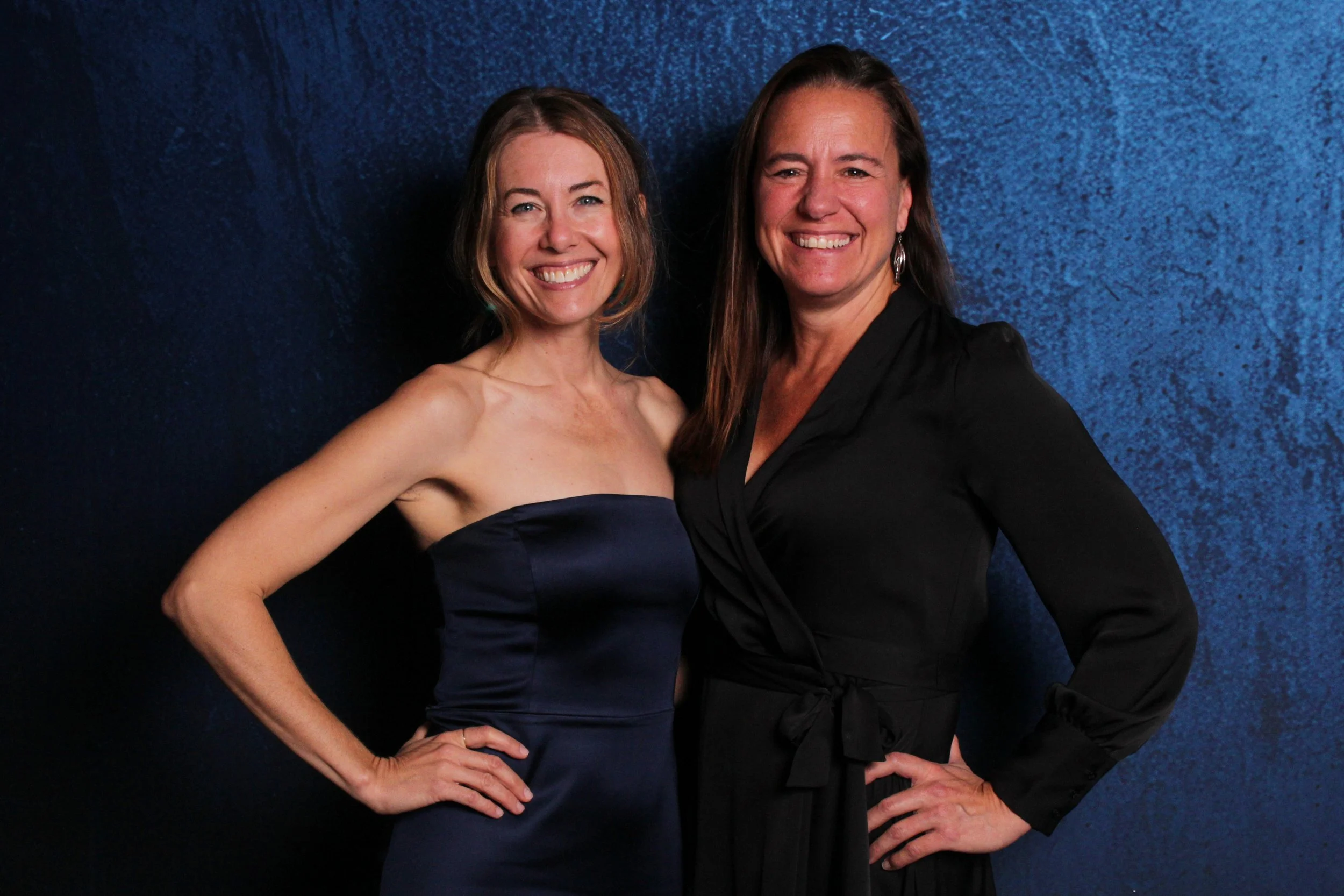 Two women smiling, standing side by side against a dark blue textured background. The woman on the left is wearing a strapless dark navy dress, and the woman on the right is wearing a black long-sleeve dress with a waist tie.