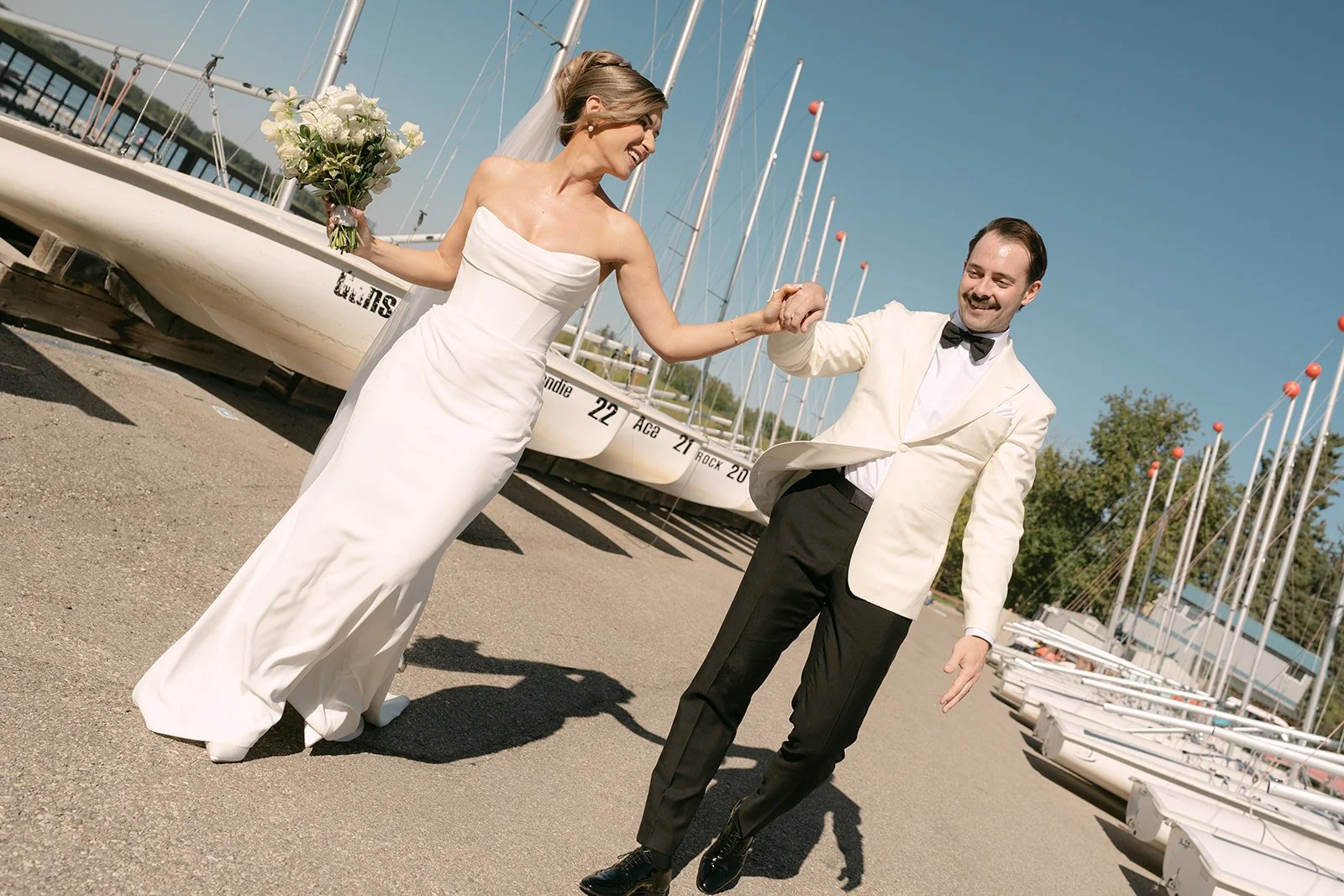 A bride and groom joyful dancing outdoors at a marina with sailboats in the background.