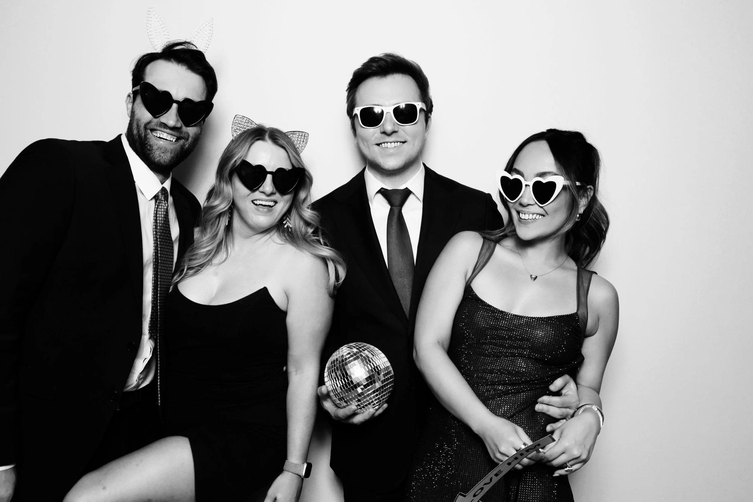 Group of four young adults dressed in formal attire, wearing playful sunglasses and accessories, smiling and posing together at a party or celebration.