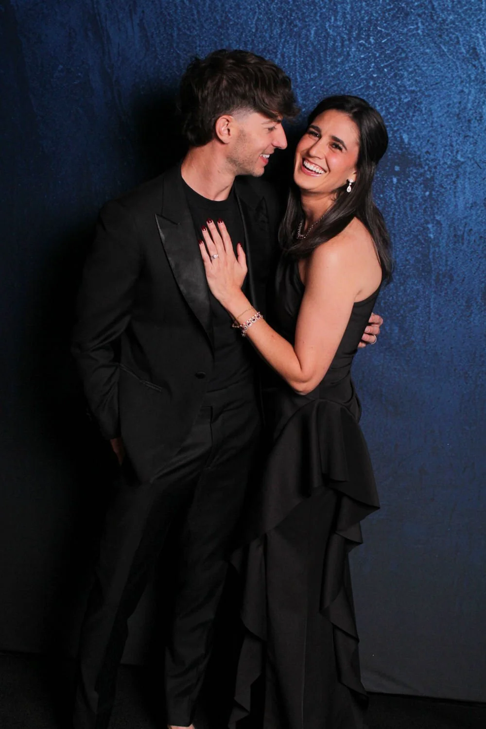 A couple dressed in formal black attire sharing a joyful moment against a dark blue textured wall, with the woman touching the man's chest and both smiling.